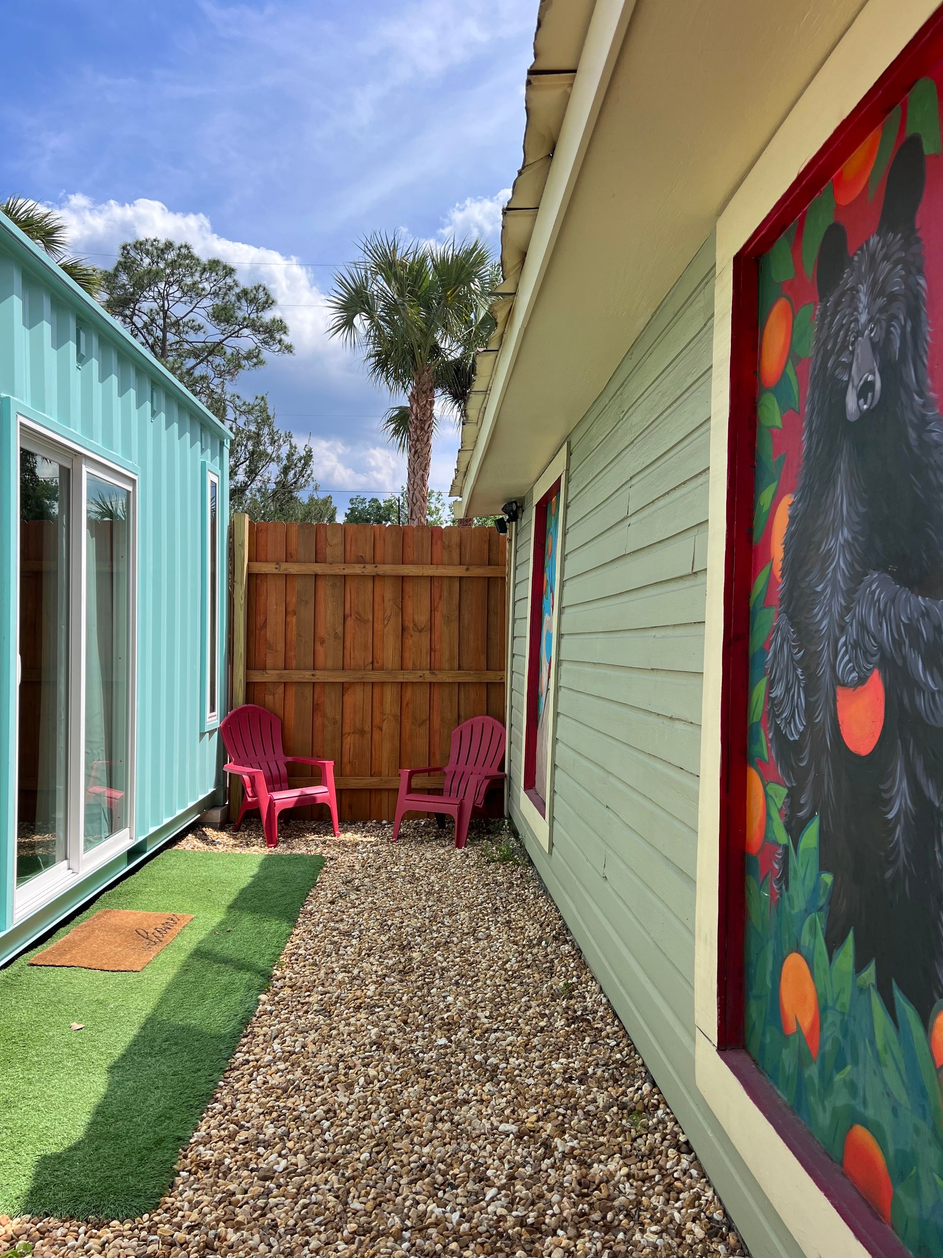 Narrow outdoor space with a turquoise container, green wall, and wooden fence. Pink chairs sit on gravel.
