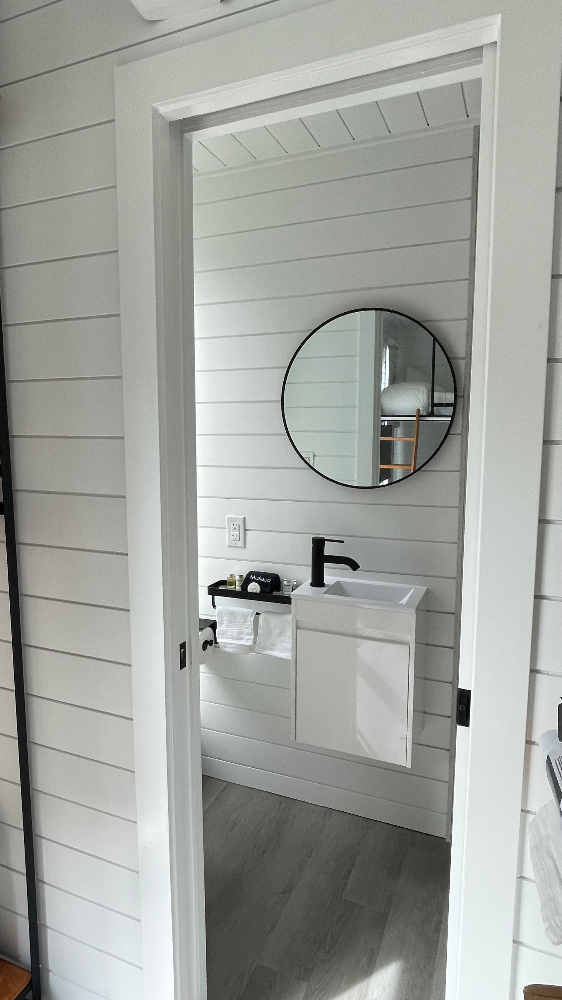 White bathroom with round mirror and wall-mounted sink seen through an open doorway.