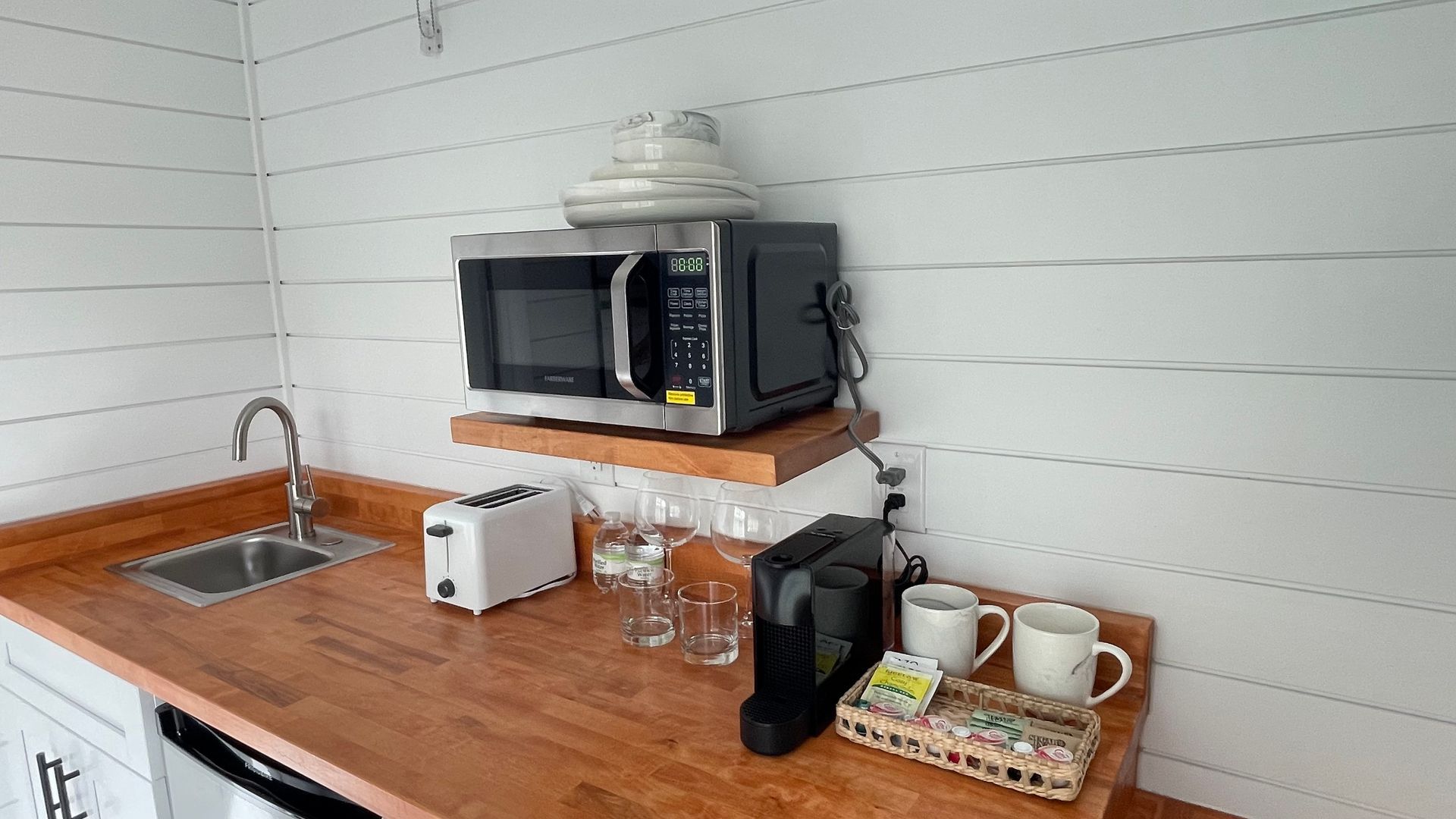 Small kitchenette with a microwave, sink, toaster, coffee maker, and mugs on a wooden countertop.