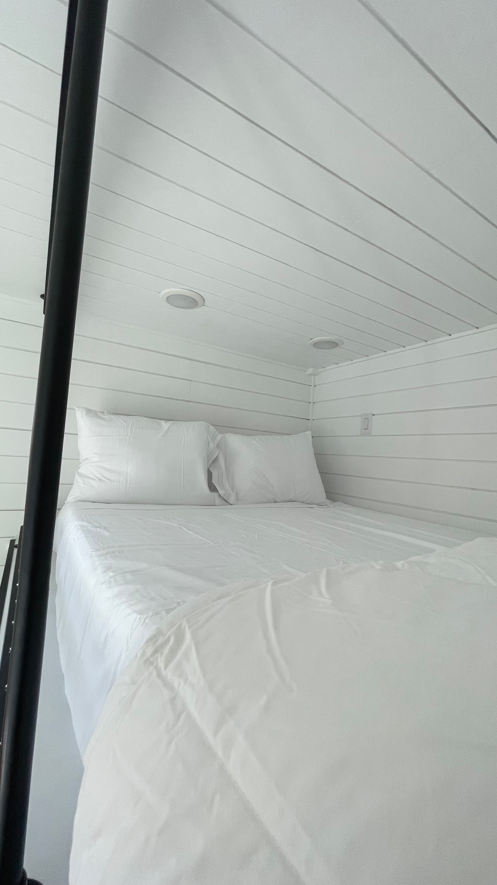 White bed with two pillows in a room with white paneled walls and ceiling.