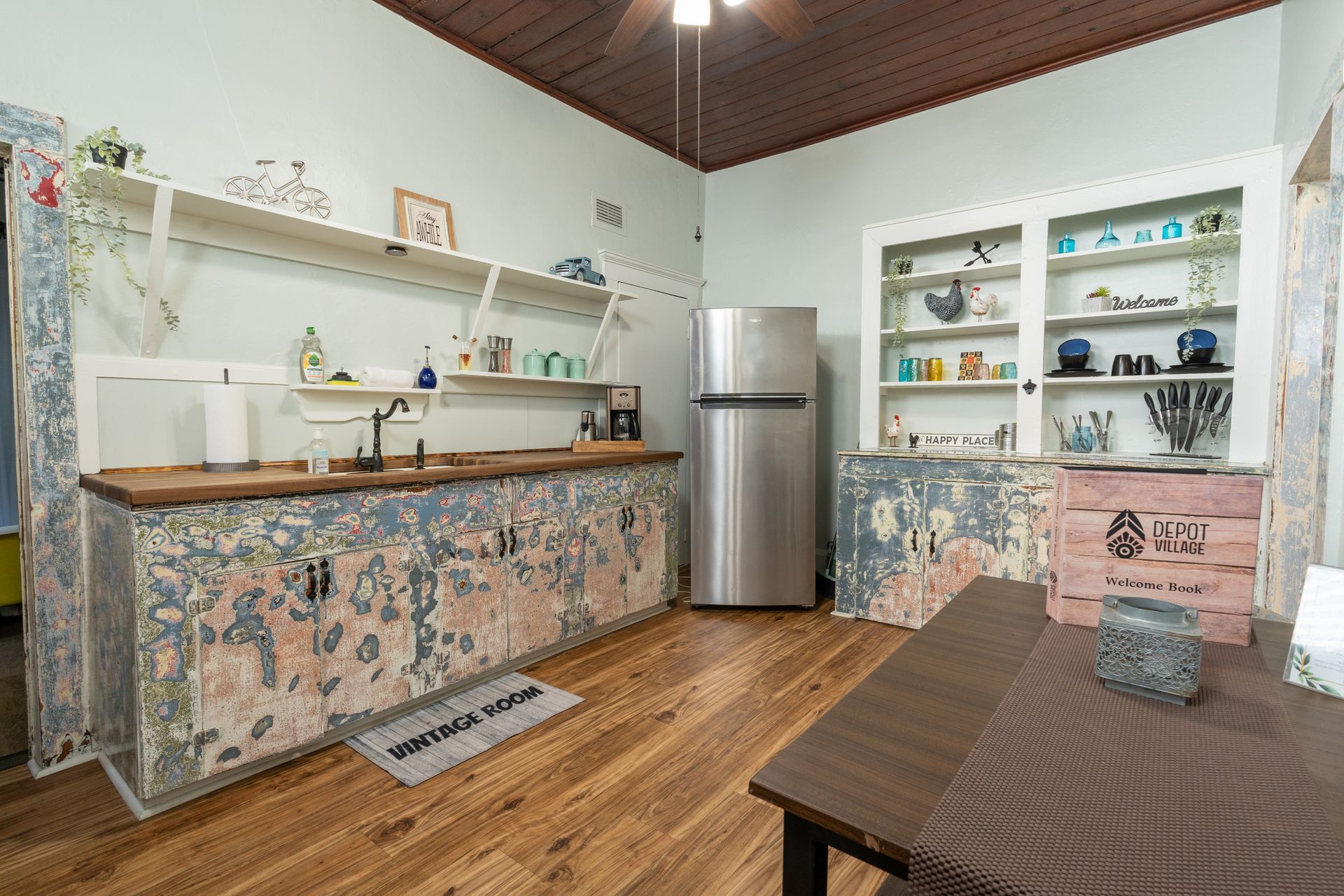Kitchen with peeling wallpaper on cabinets, stainless steel fridge, wooden countertops, shelves, and wood floors.