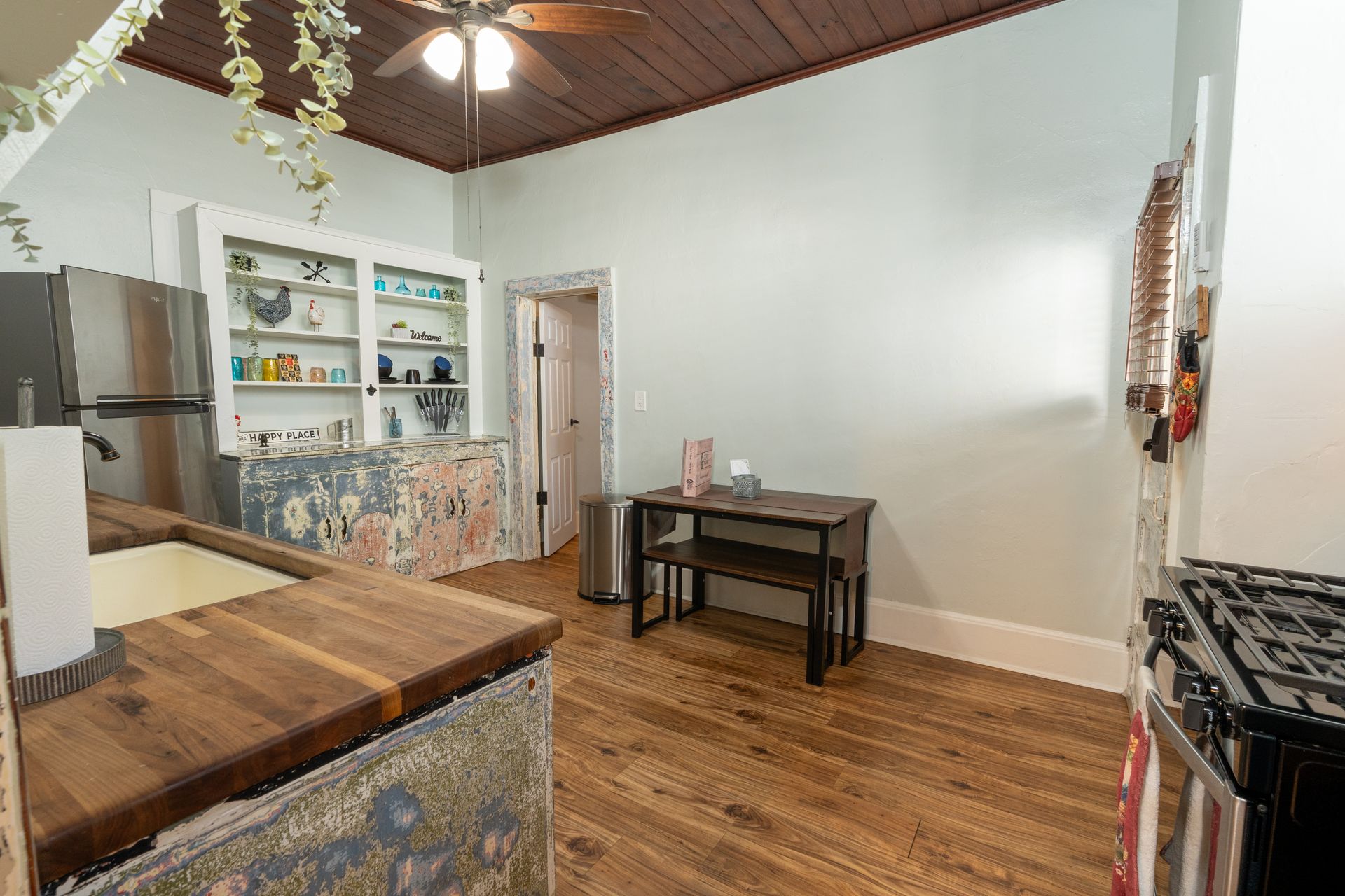 Kitchen with wooden floors, countertops, and dark wooden table. White shelves, floral design, stainless steel appliances.