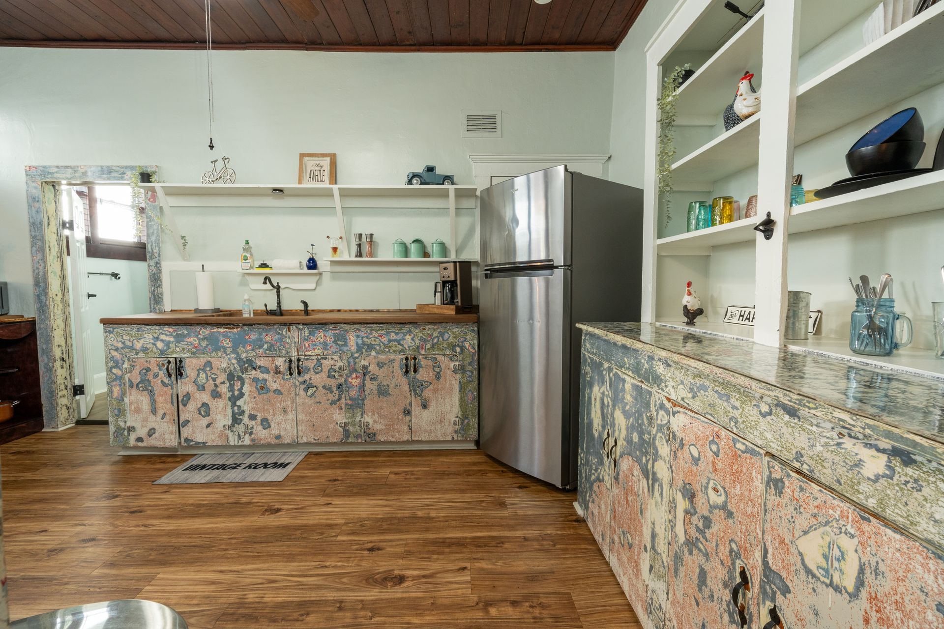 Kitchen with distressed cabinets, open shelves, stainless steel refrigerator, and wood flooring.
