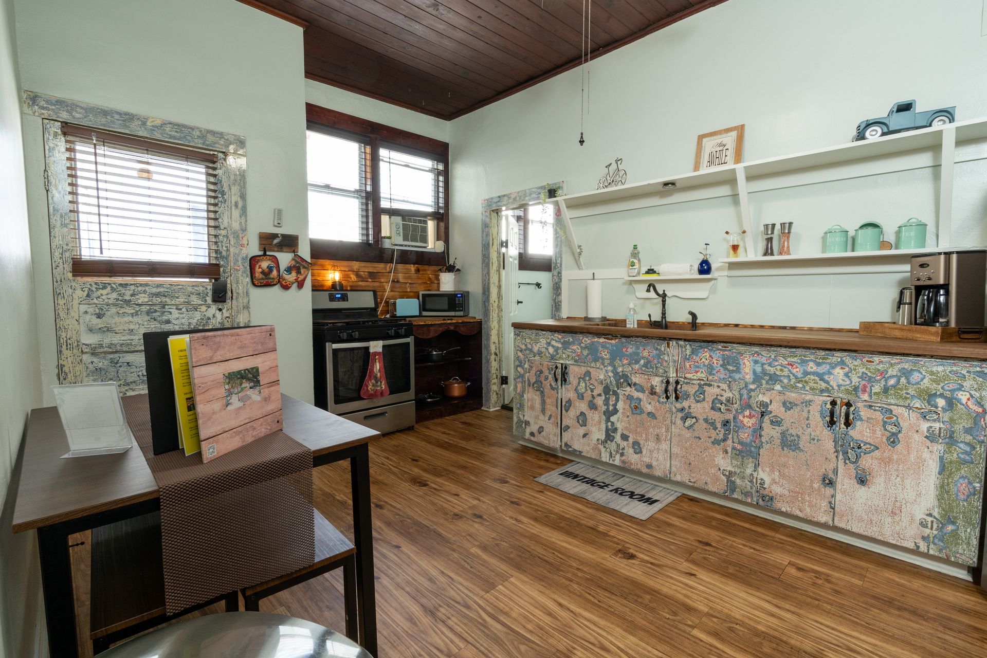Kitchen with floral-patterned cabinets, wooden floor, and open shelving. Includes stove, microwave, and small dining table.