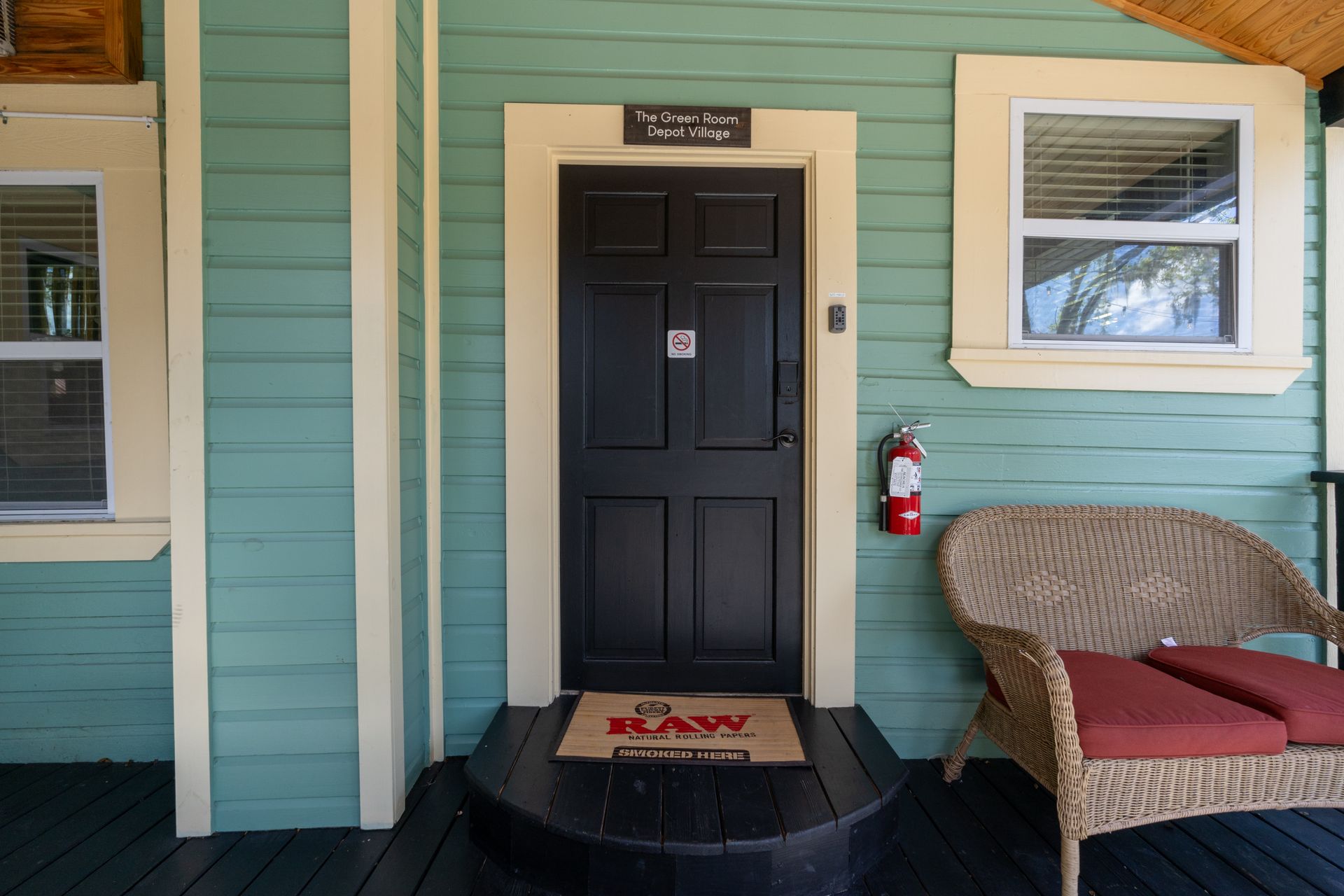 Exterior of a teal building with a black door, doormat, wicker chair, and fire extinguisher.