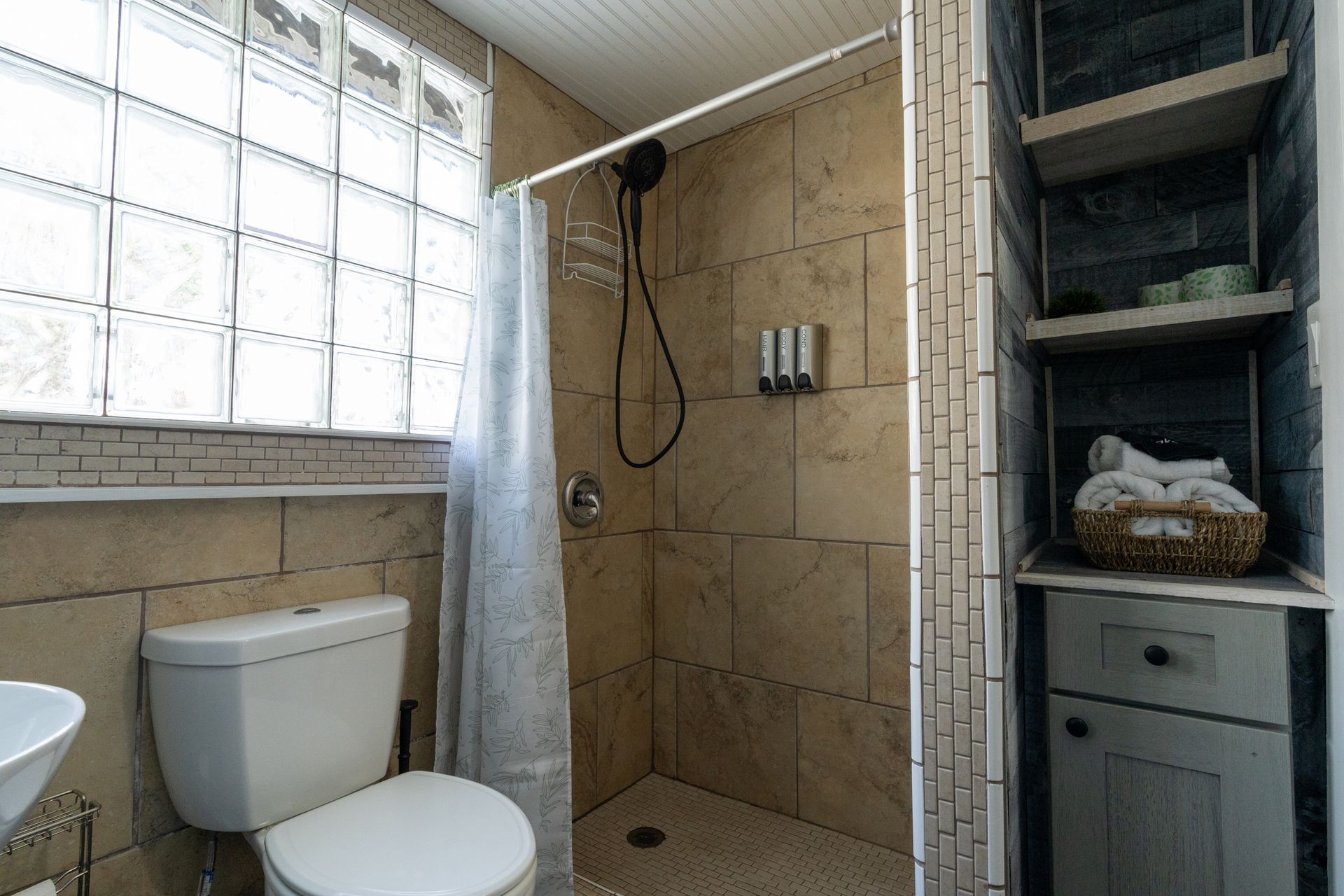 Bathroom with a shower, toilet, and shelving unit. Beige tiles, glass block window, gray curtain, and white sink.