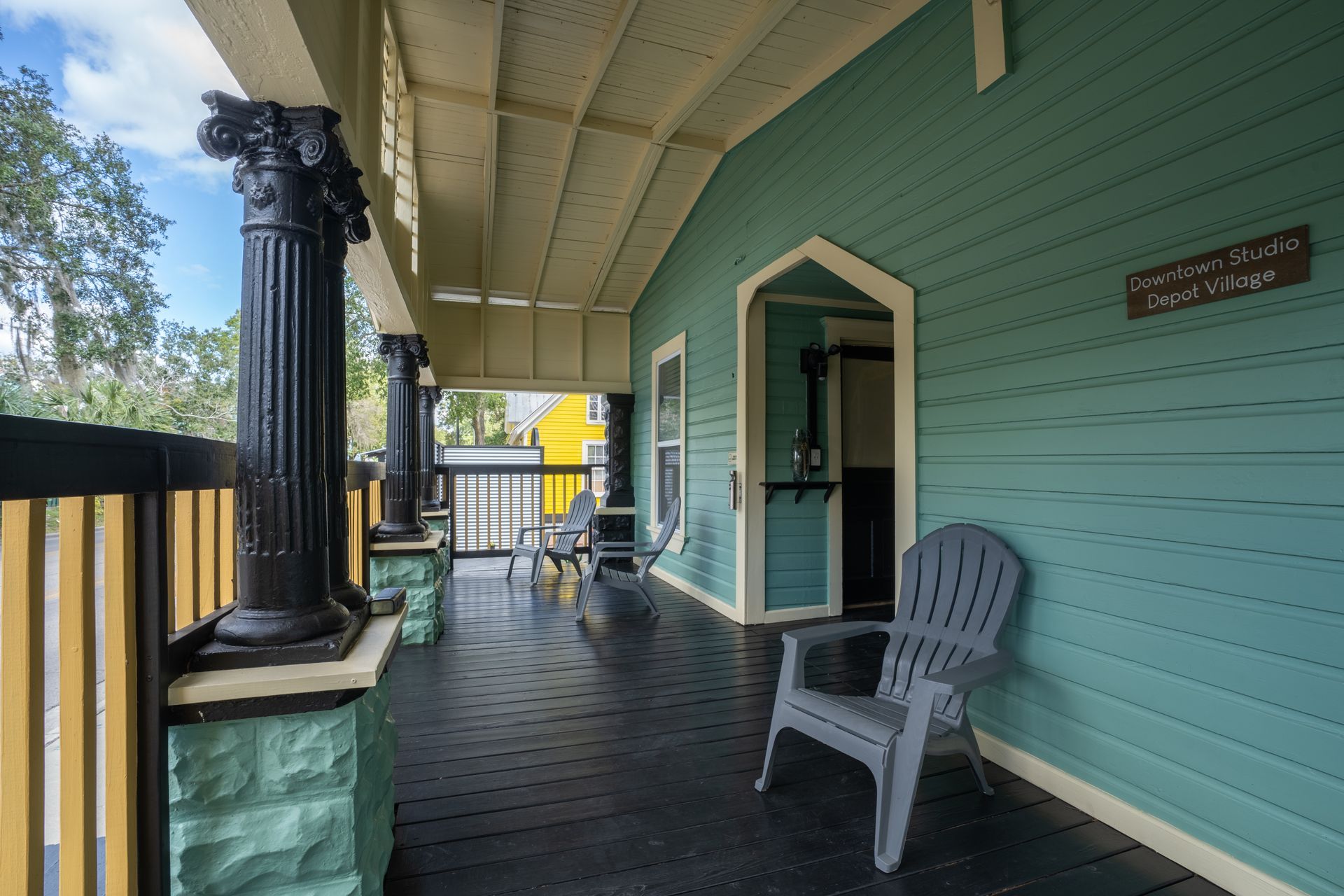 A teal-painted porch with black columns and a gray chair. Open doorway with chairs visible in the background.
