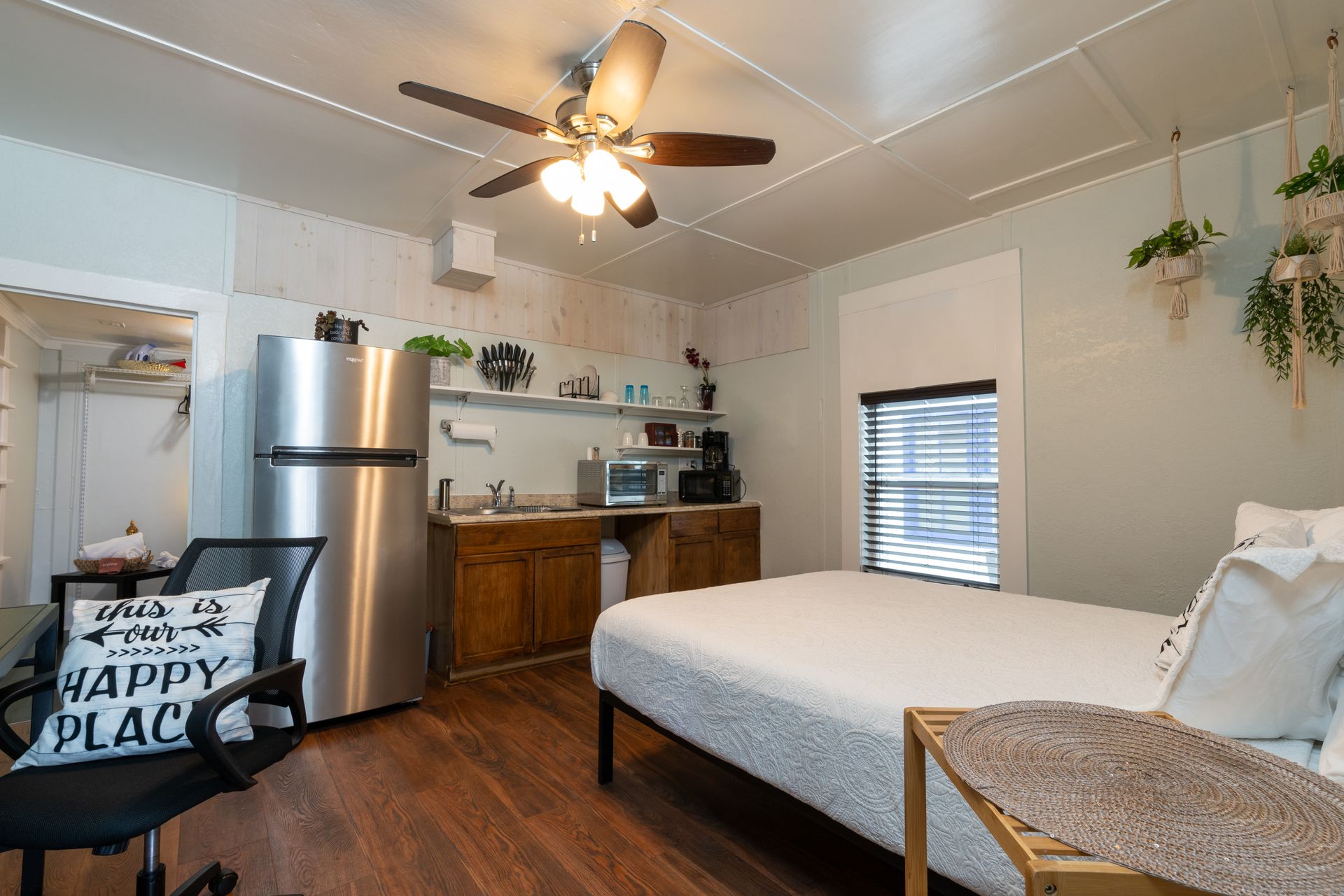 Studio apartment with bed, small kitchen, desk chair, and refrigerator.
