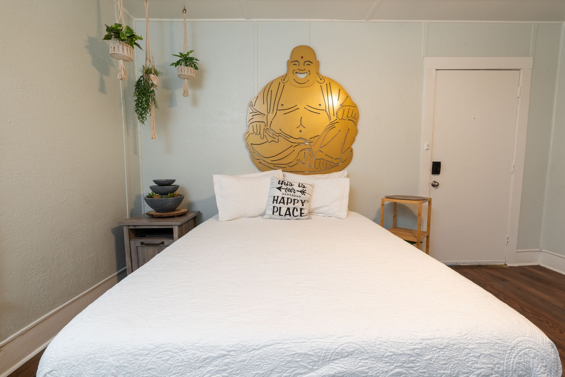Bedroom with white bedding, Buddha headboard, hanging plants, and a small side table.