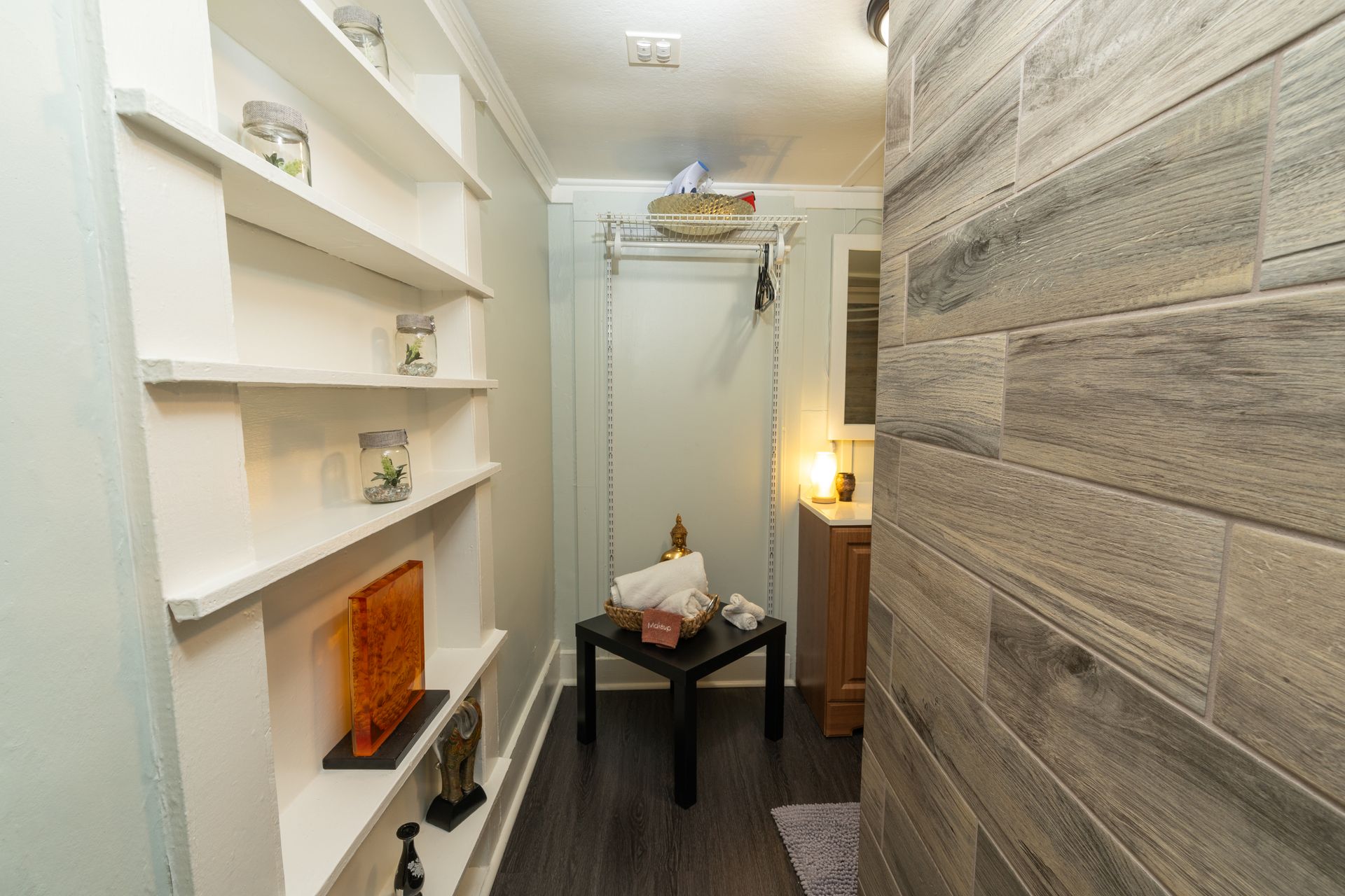 Narrow hallway with built-in shelves on left, gray tile wall on right, and black table with decor in the center.