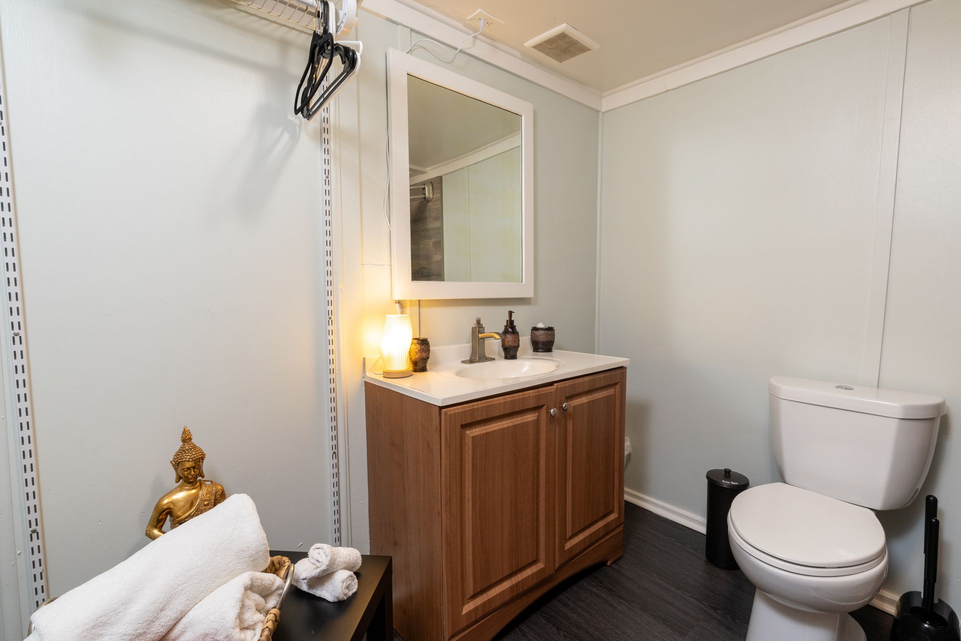 Bathroom with vanity, mirror, toilet, and gold Buddha statue. Light blue walls, wooden cabinet, white towels.