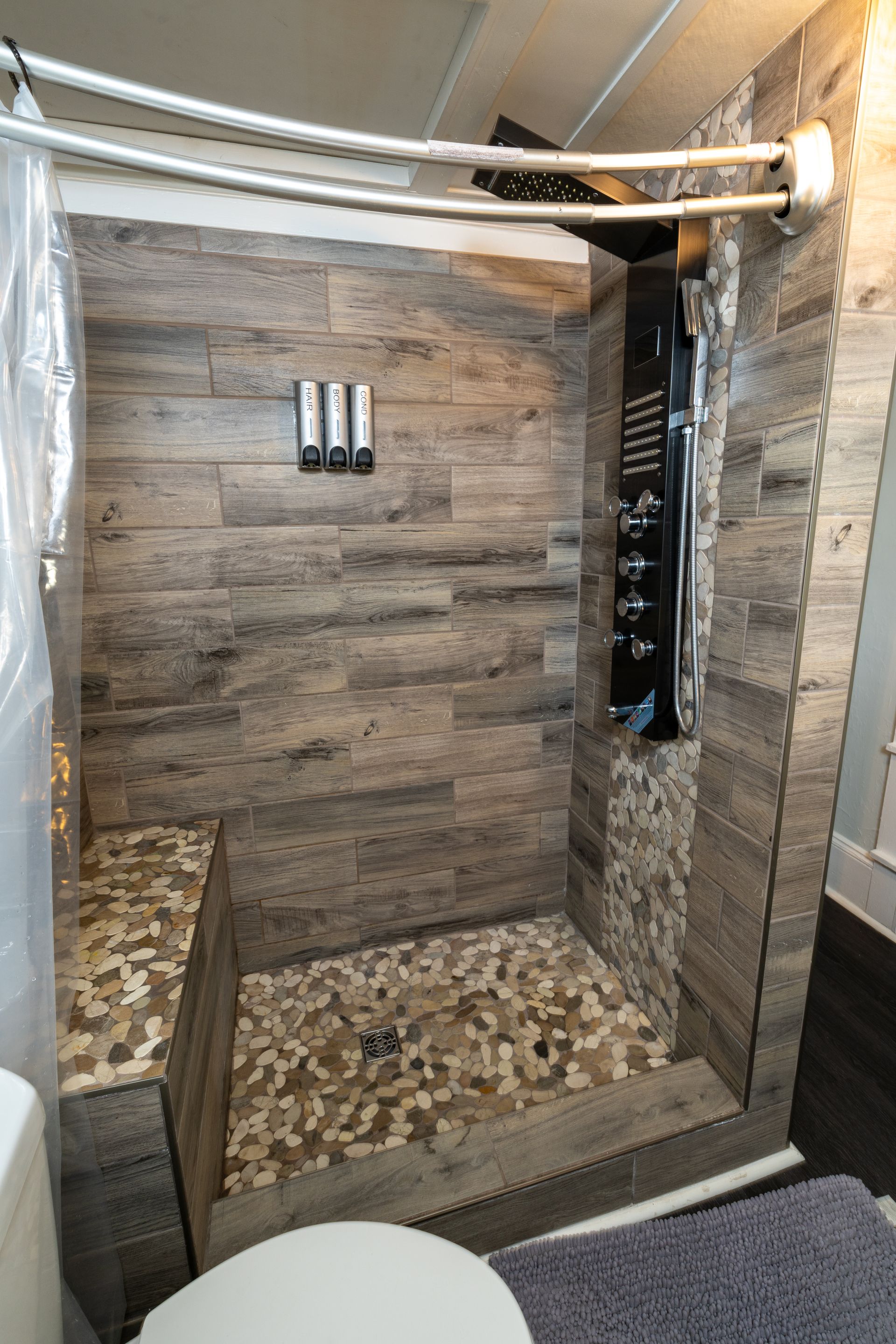 Small walk-in shower with wood-look tile, pebble floor, and black shower panel; a shower curtain is to the left.