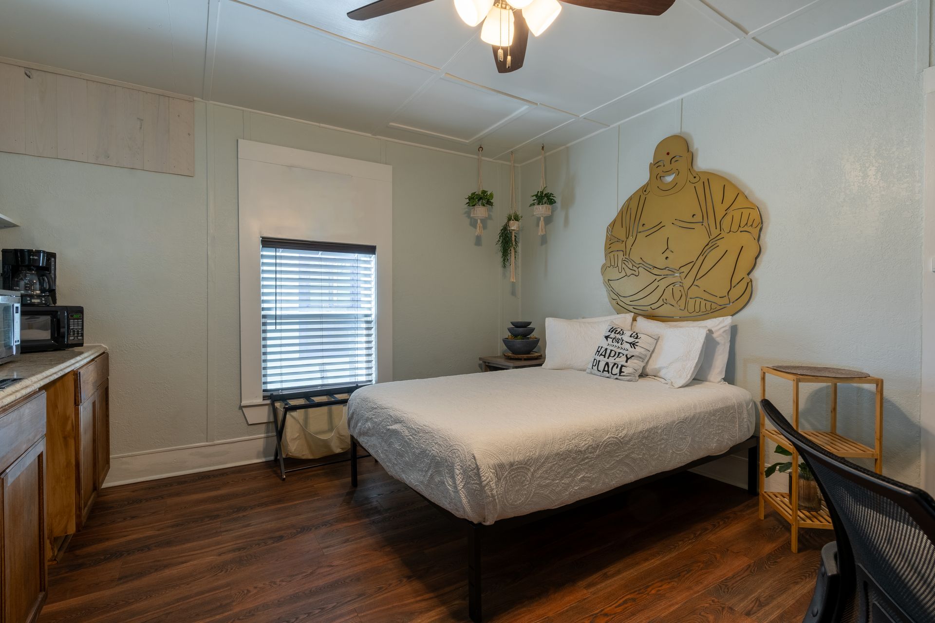 Small bedroom with bed, window, side table, and kitchenette. Wooden floors and walls are light green.