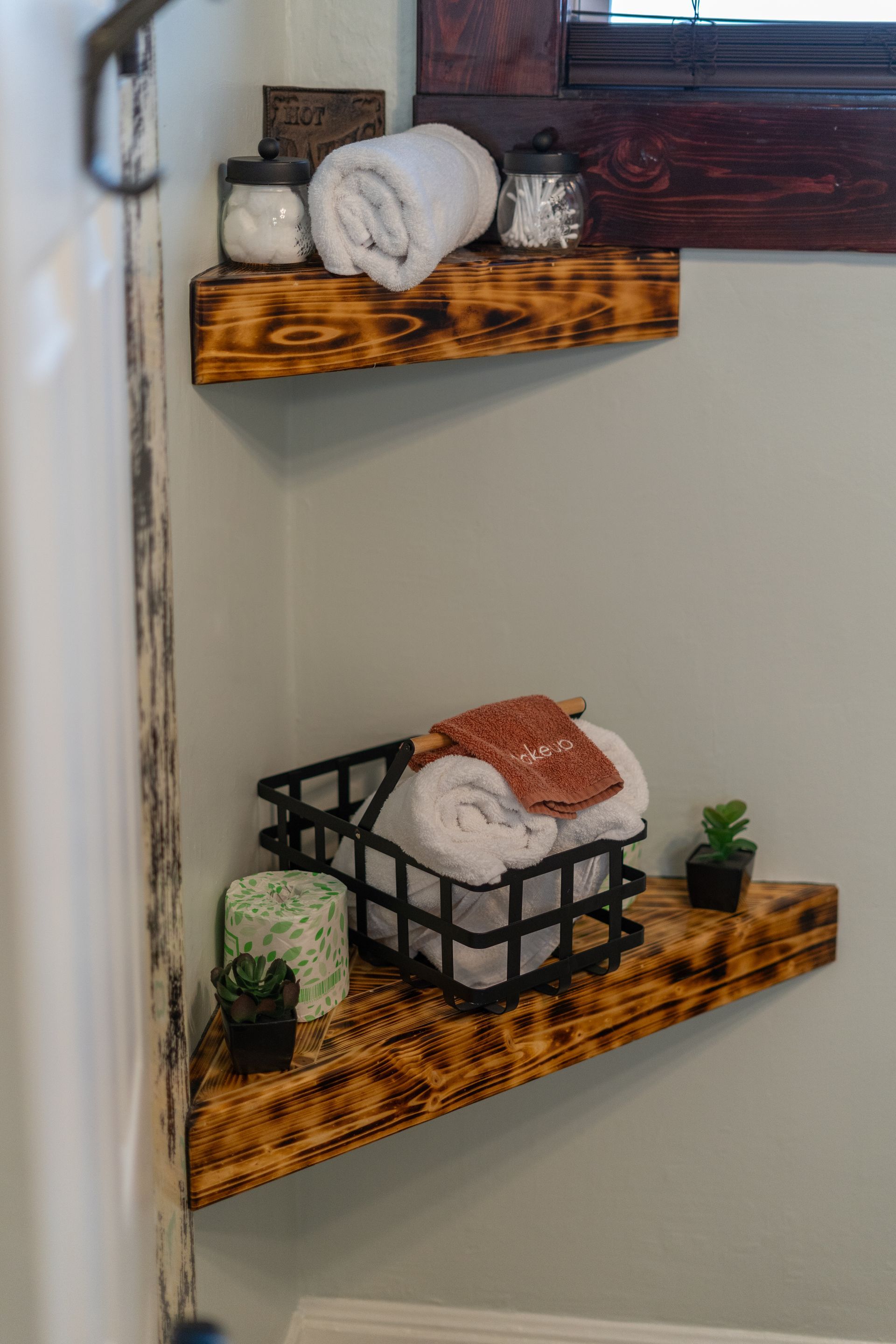 Corner shelves in a bathroom hold towels, jars, and small plants. Burnished wood against a light wall.