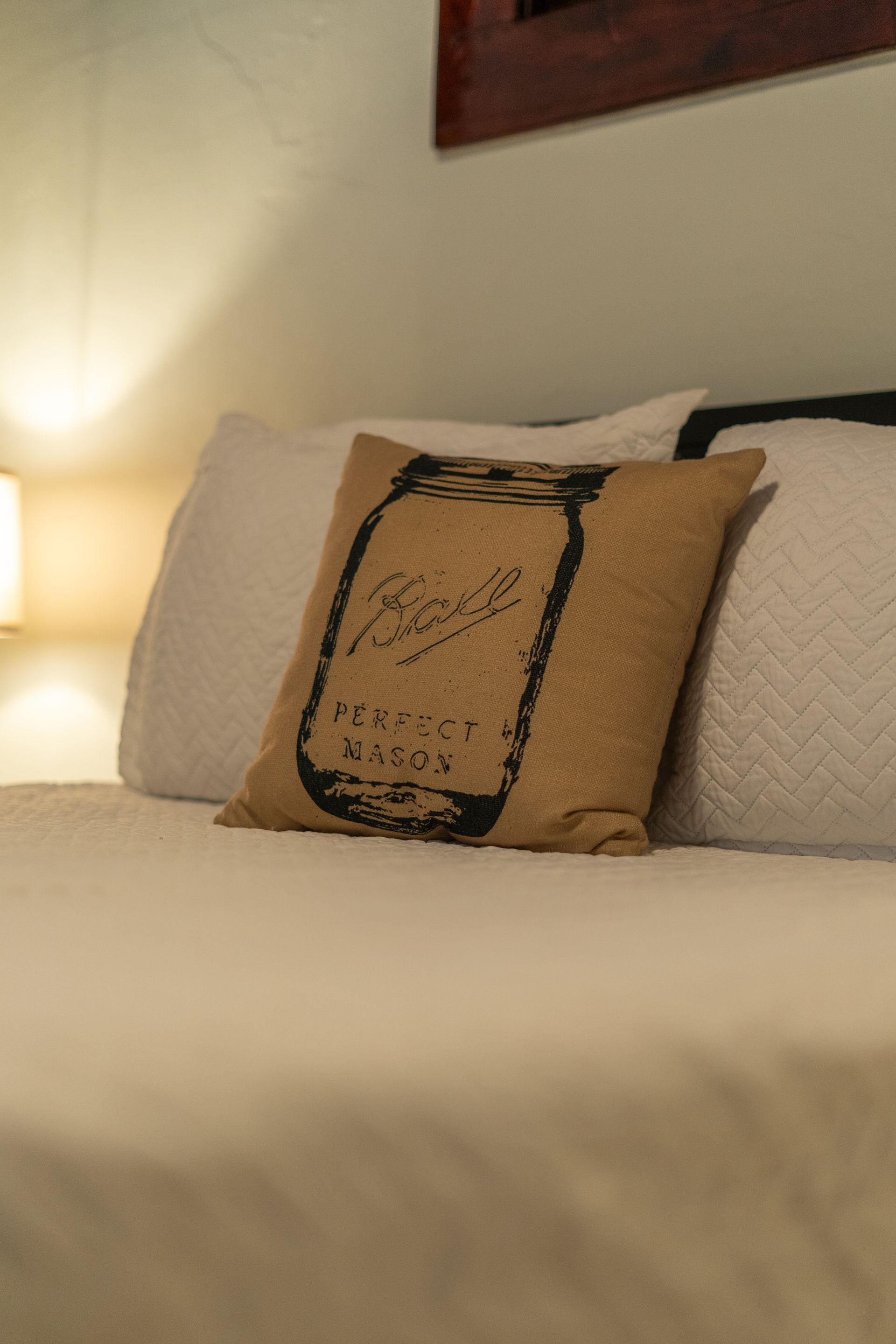 Bed with white bedding and a burlap pillow with a printed jar design.