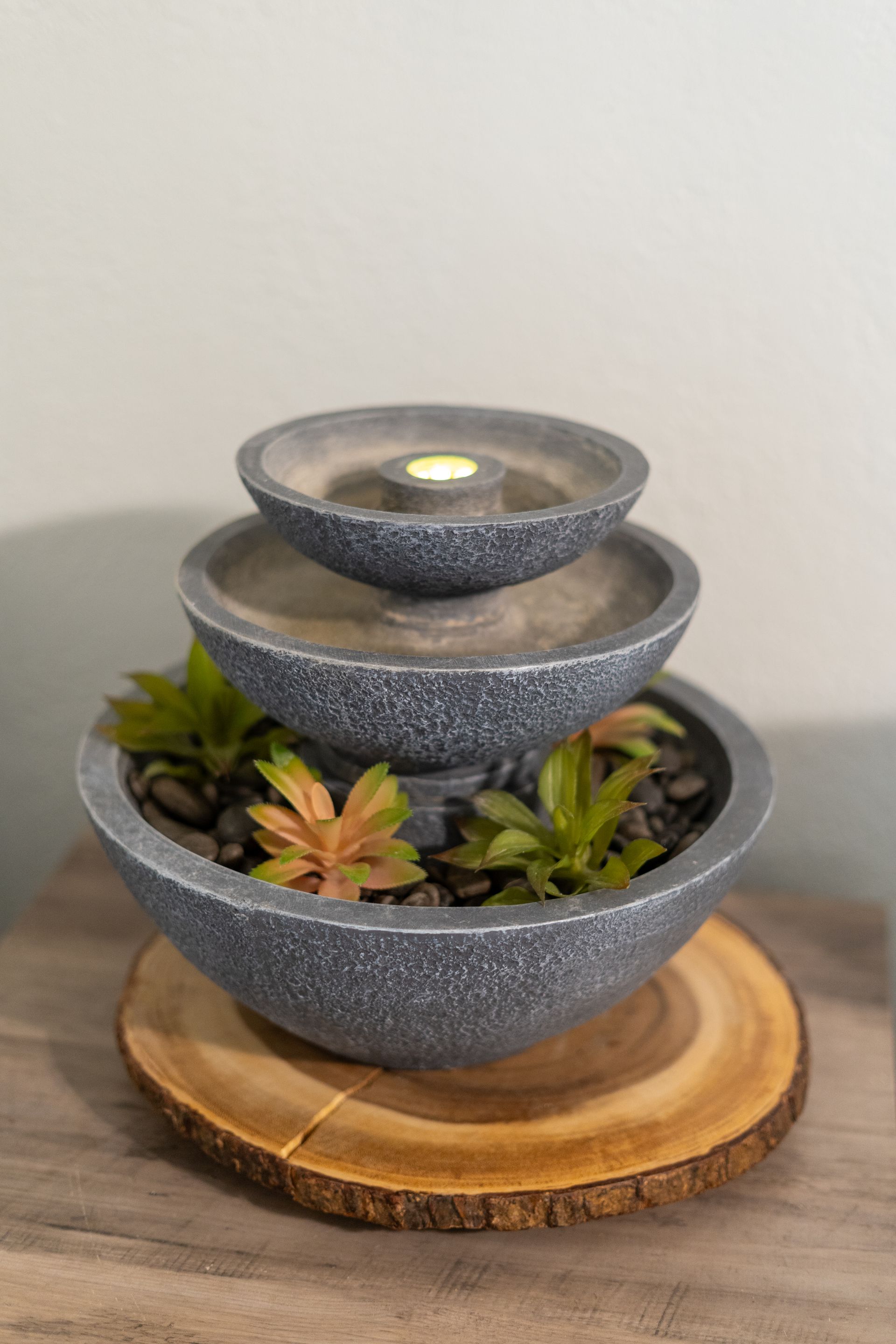 Three-tiered water fountain with succulents, black stones, and a candle, resting on a wooden slice.