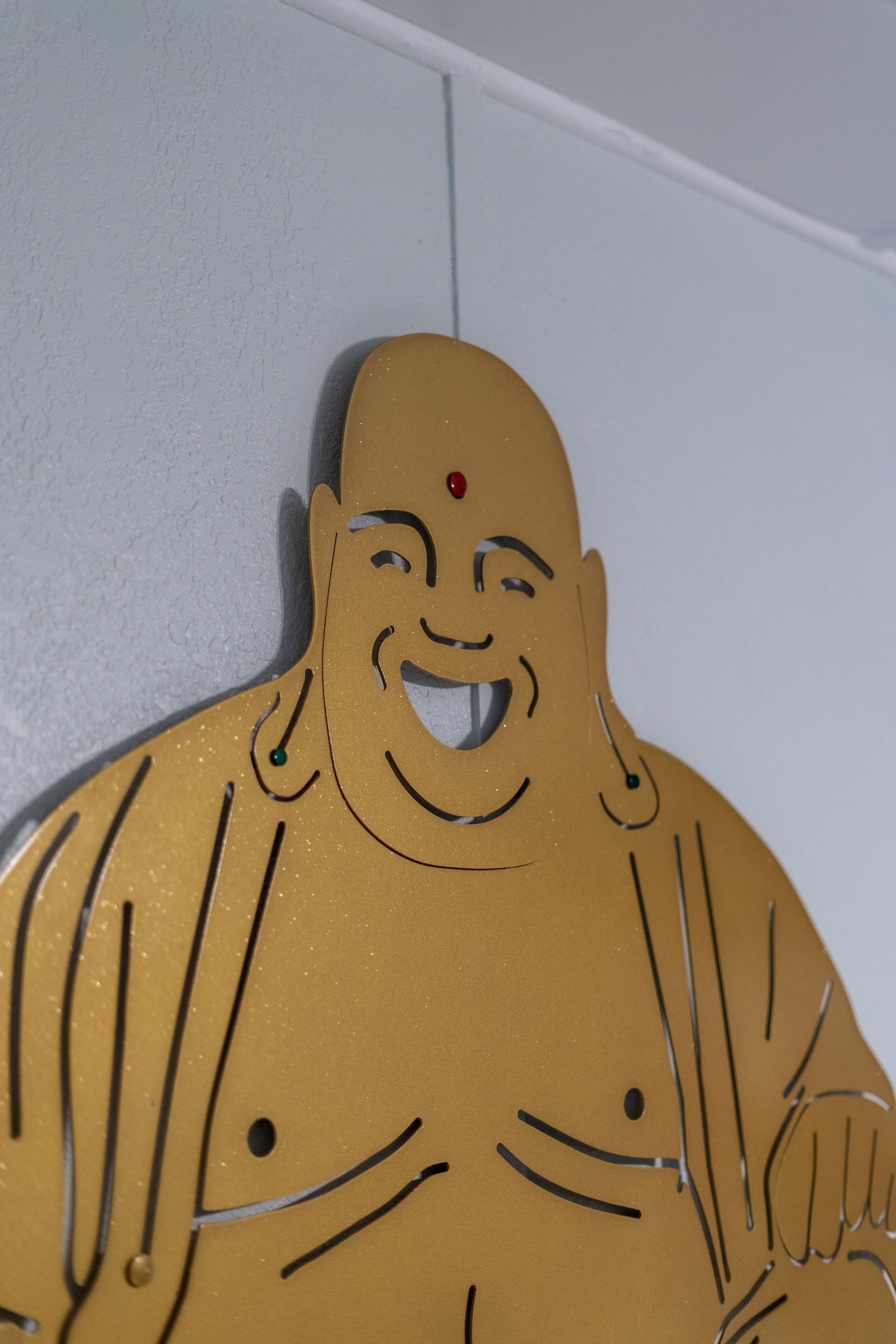 Golden, smiling Buddha wall art with red dot, hanging on a light blue wall.