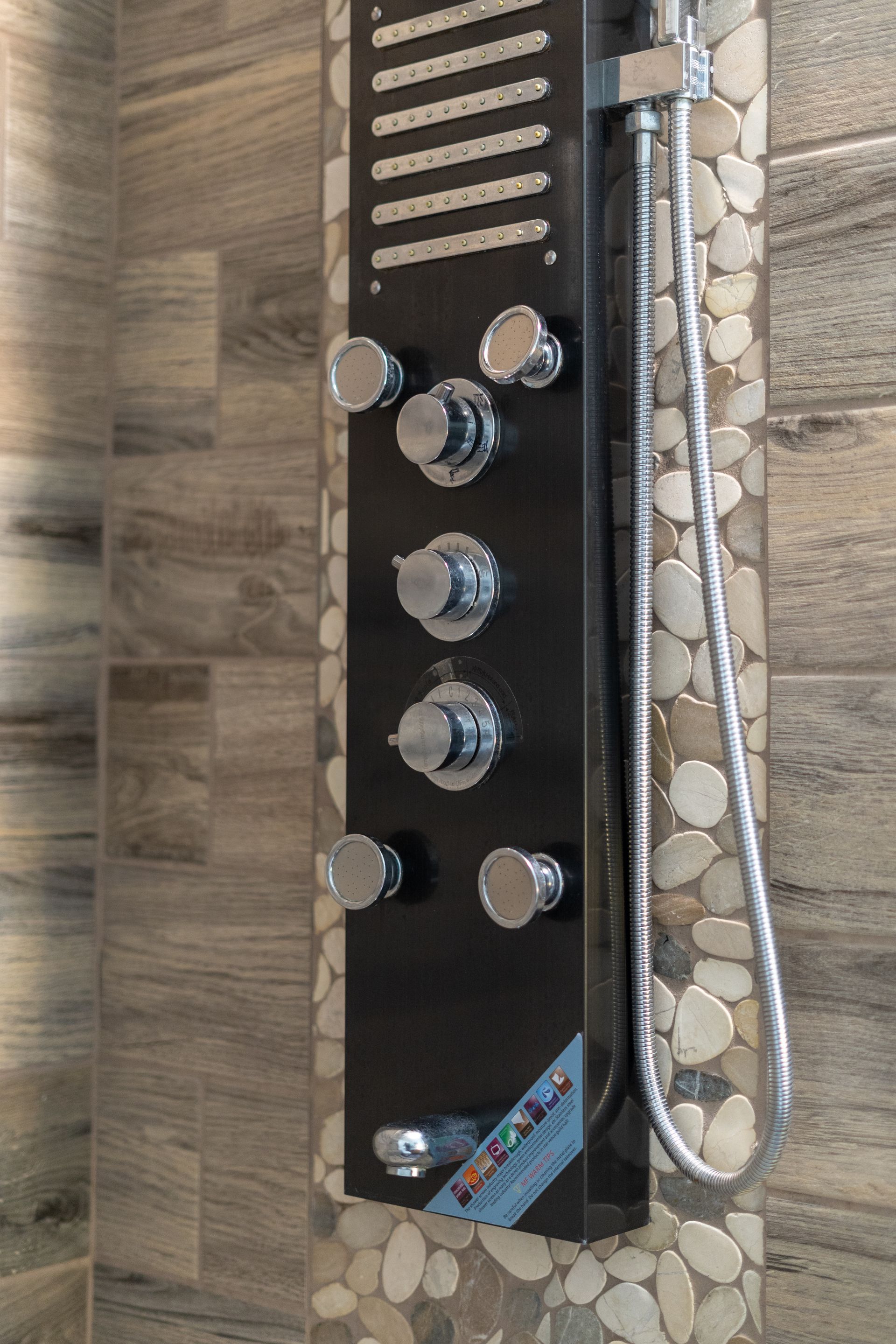 Black shower panel with chrome knobs and showerhead, mounted on a stone-tiled wall.