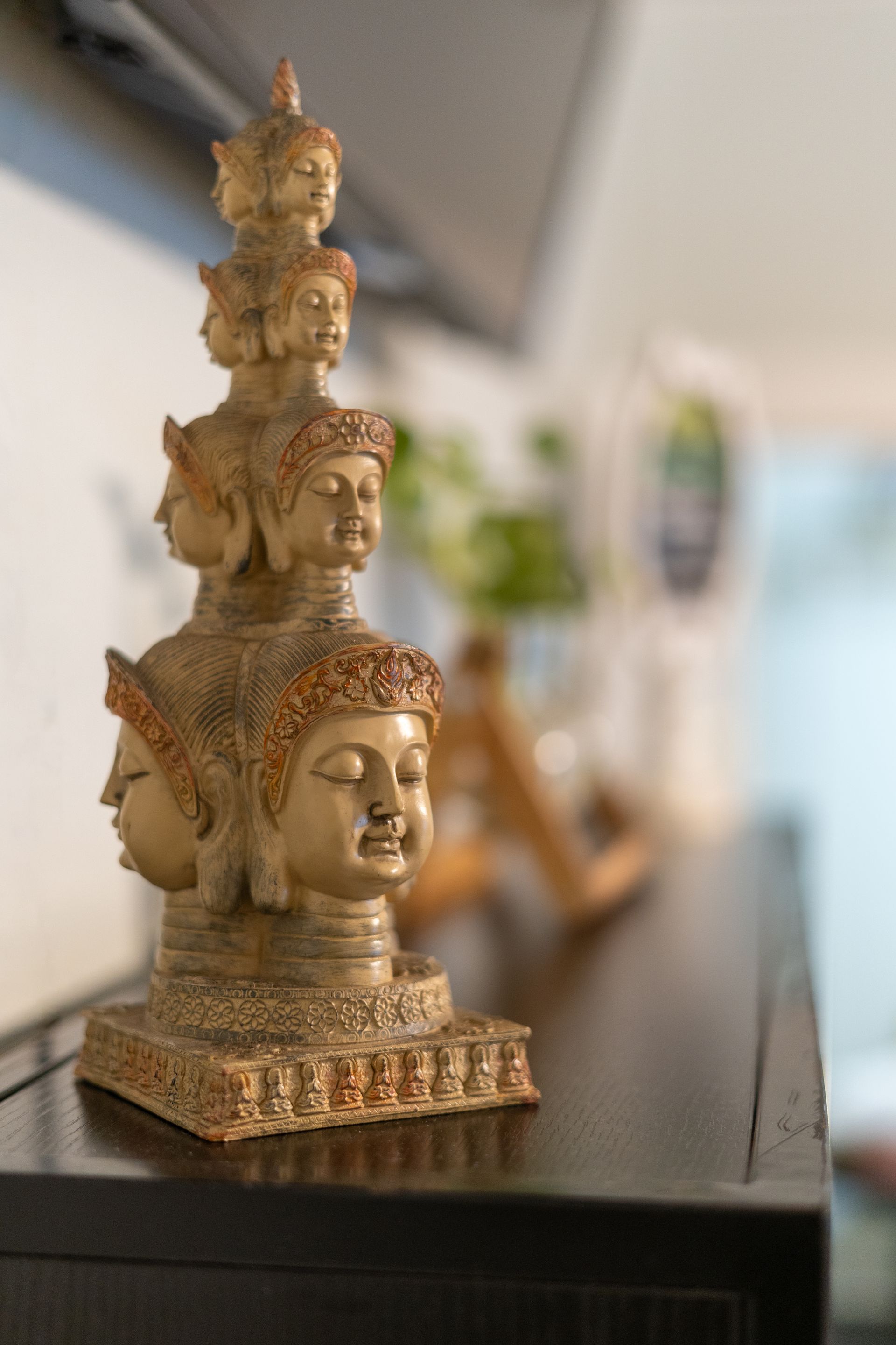 Tan-colored Buddha statue with multiple faces, on a dark surface, blurred background.