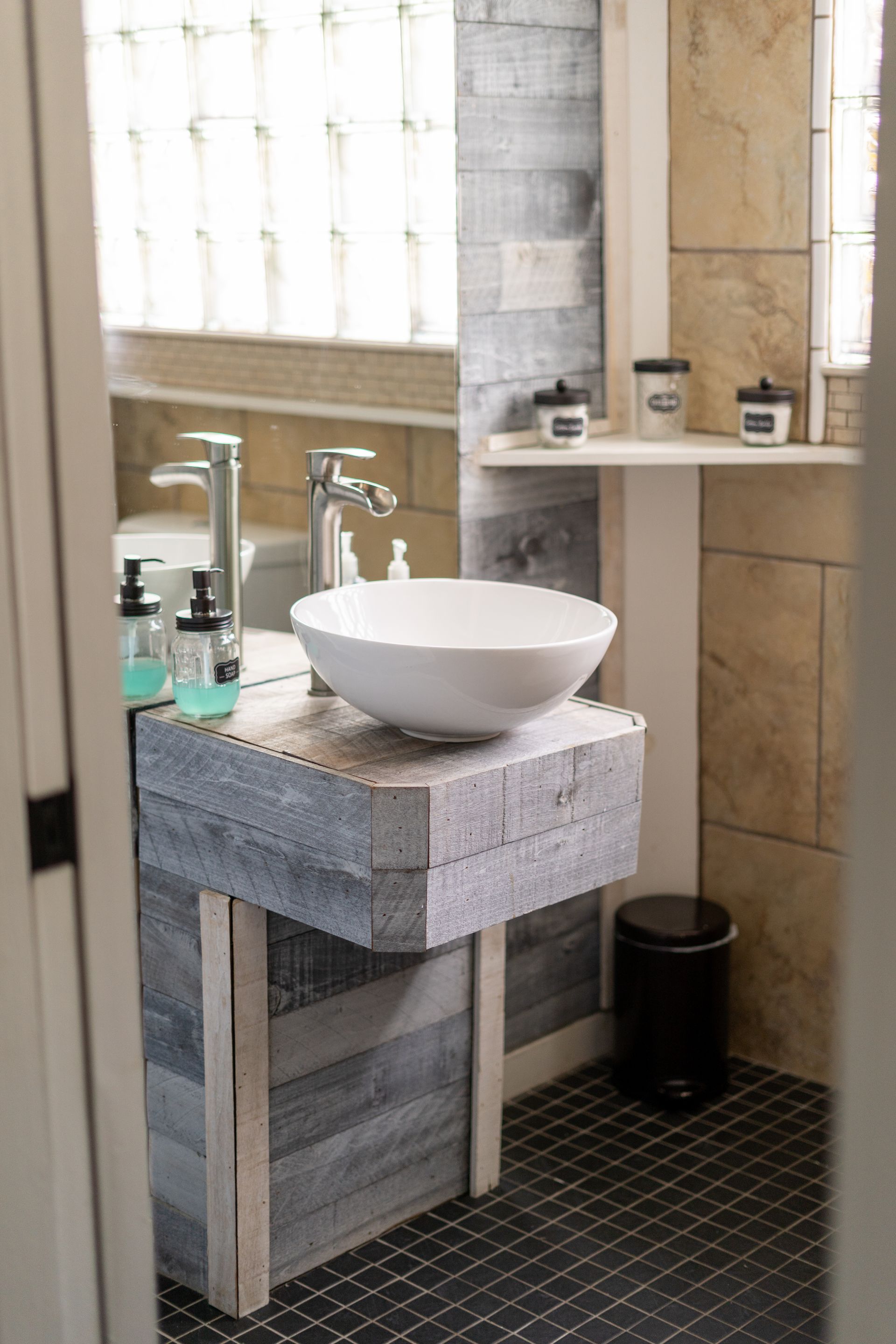 A bathroom with a vessel sink on a gray wooden stand, black trash can, and tiled walls.