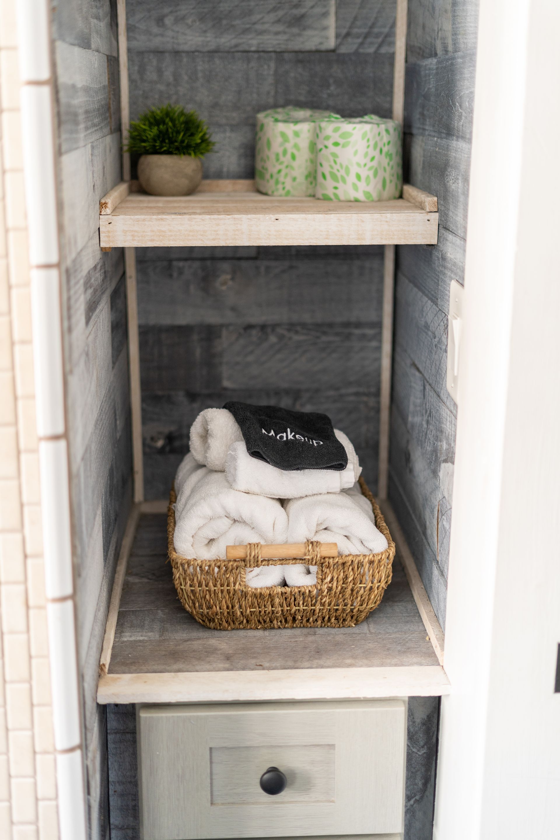 Built-in bathroom shelving with towels, toilet paper, and a small plant. The walls are gray wood paneling.