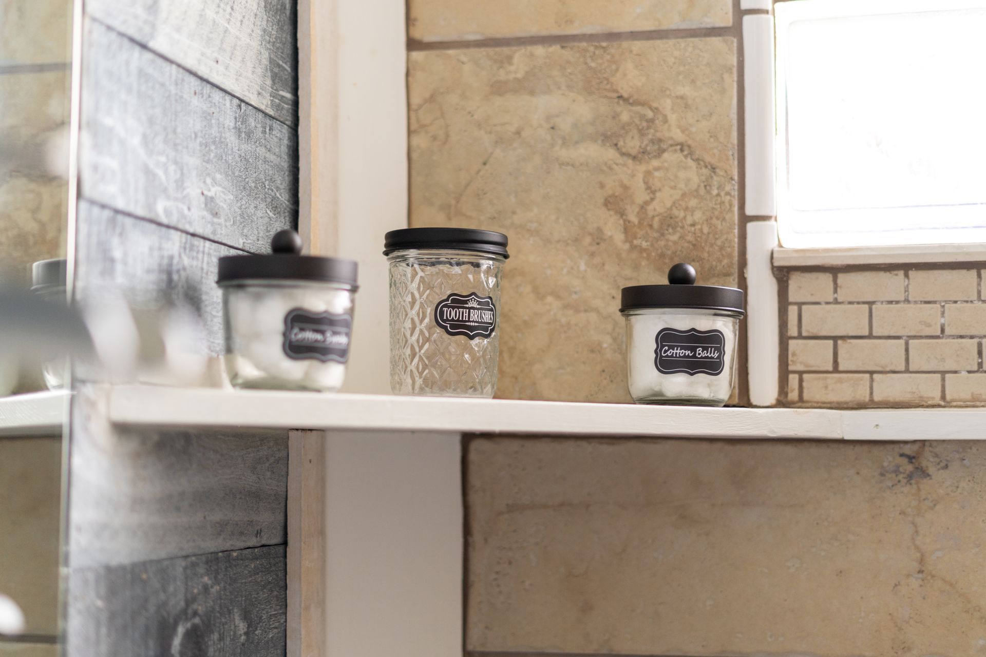 Three jars on a white shelf in a bathroom; two filled with cotton balls, one empty, against a tile backdrop.