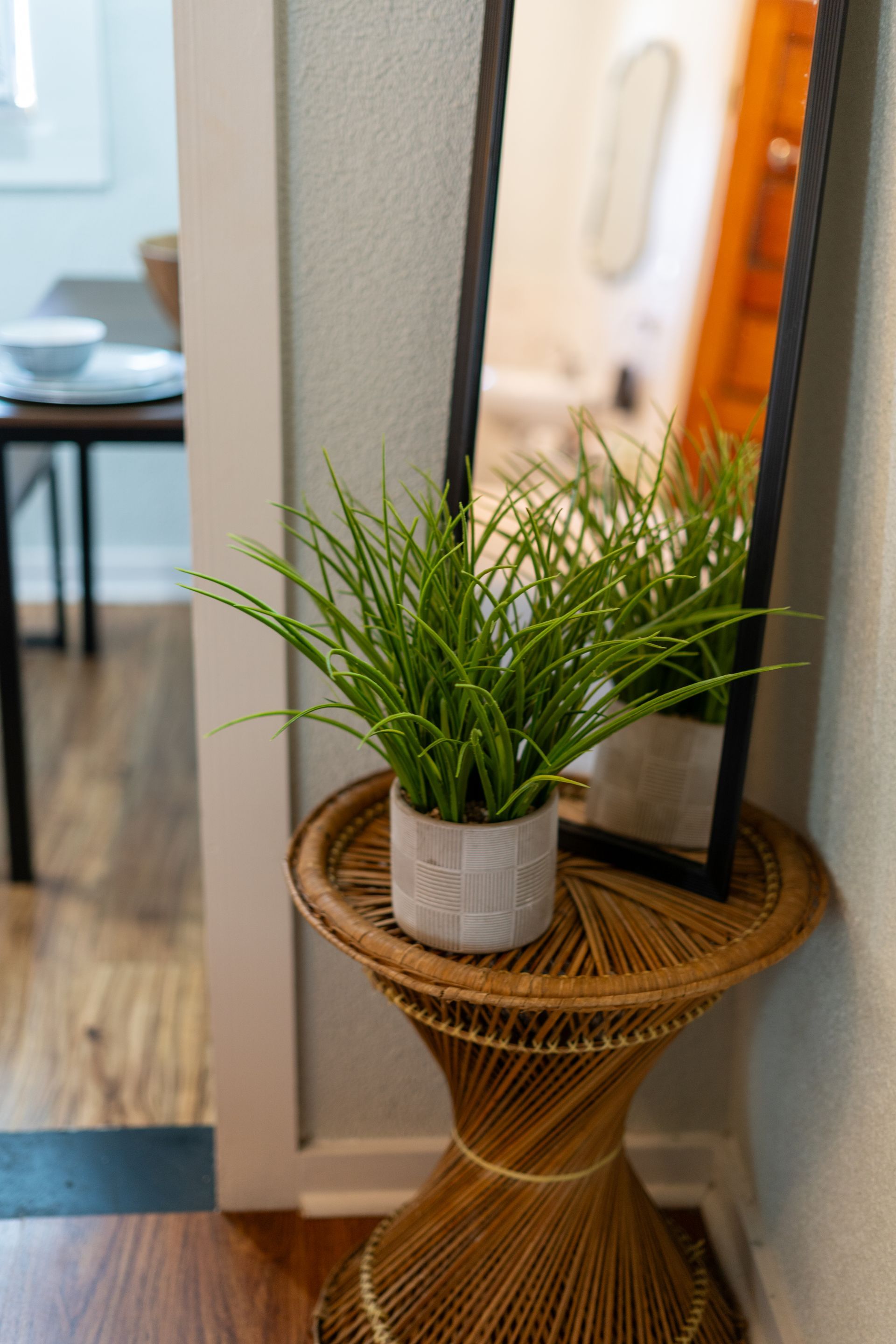A potted green plant on a woven side table next to a full-length mirror, set in a corner of a room.
