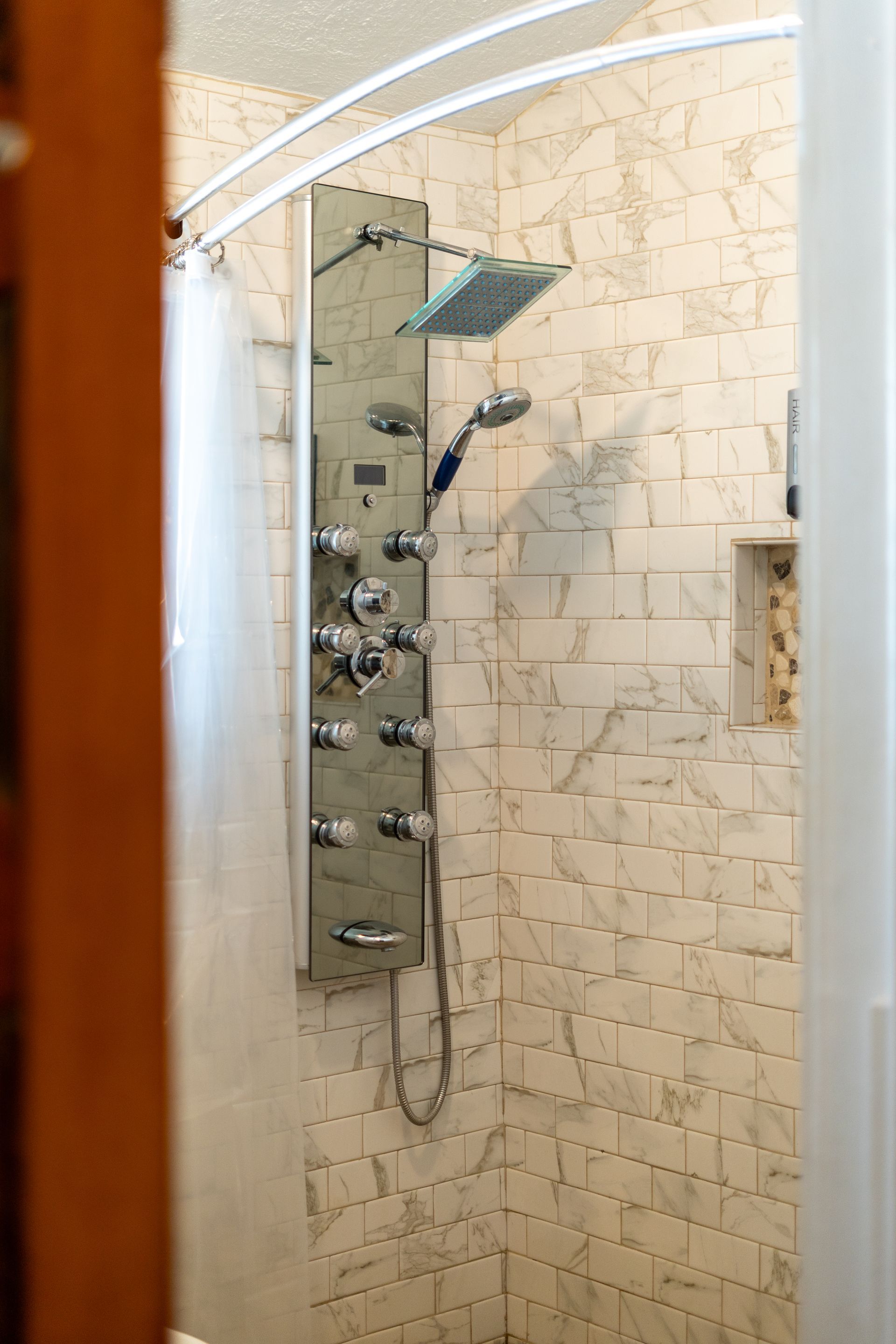 Shower stall with white marble-look tiles, chrome shower panel with multiple controls, and a curved shower rod.