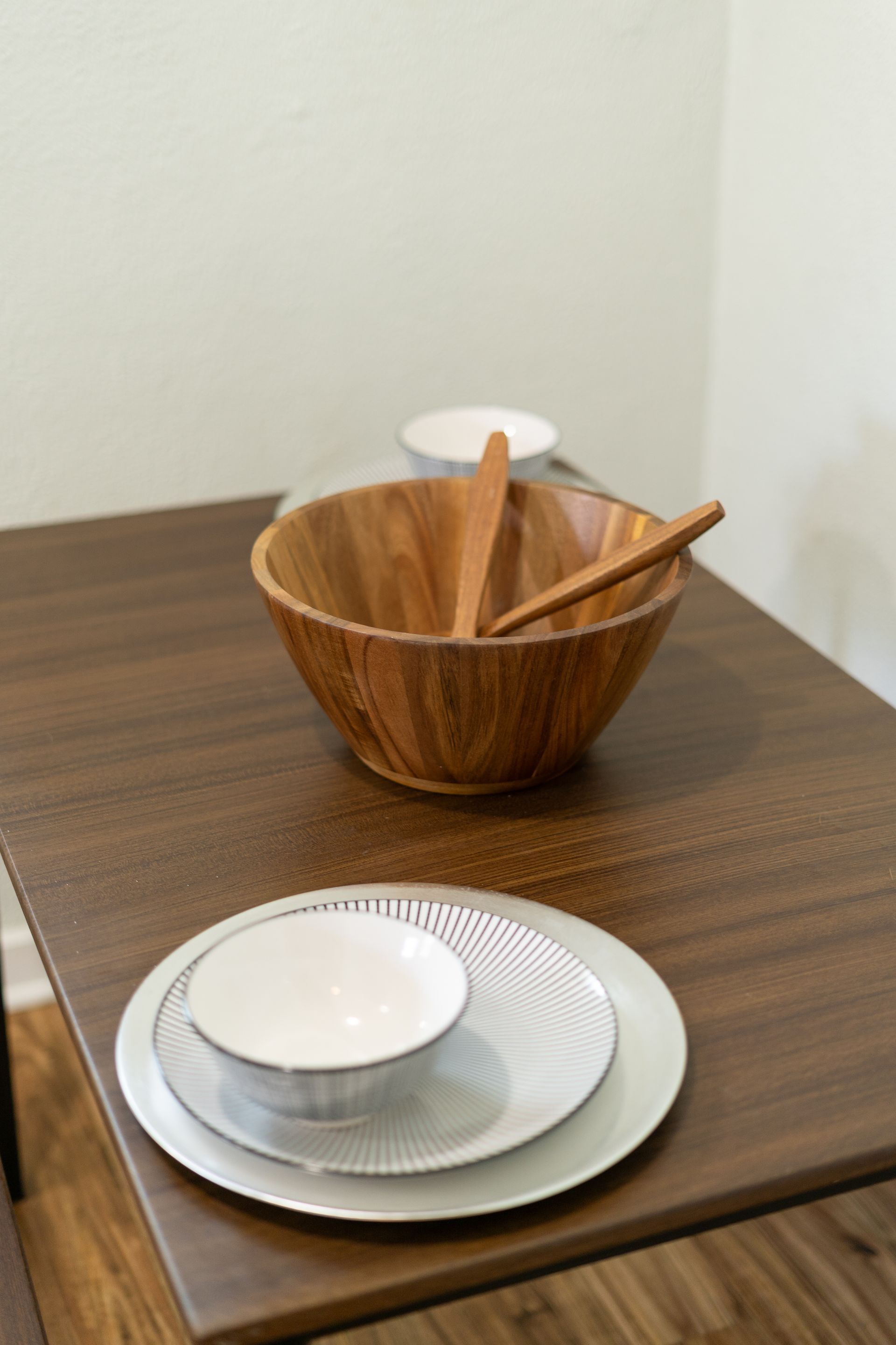 Wooden salad bowl with utensils and a set of plates on a dark brown table.