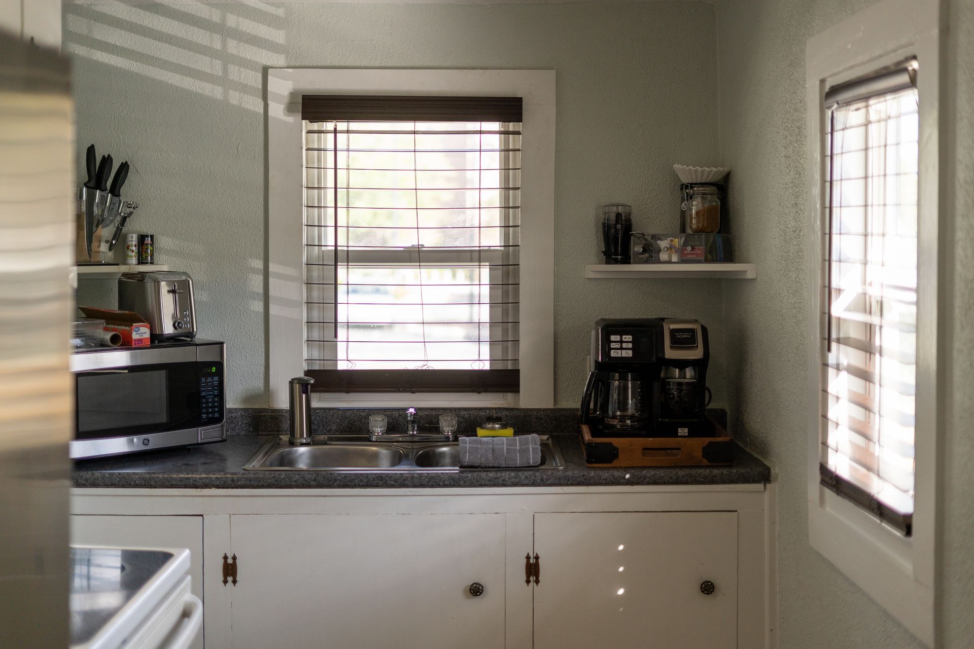 Kitchen with sink, microwave, coffee maker, and two windows with blinds. Light streams through windows.