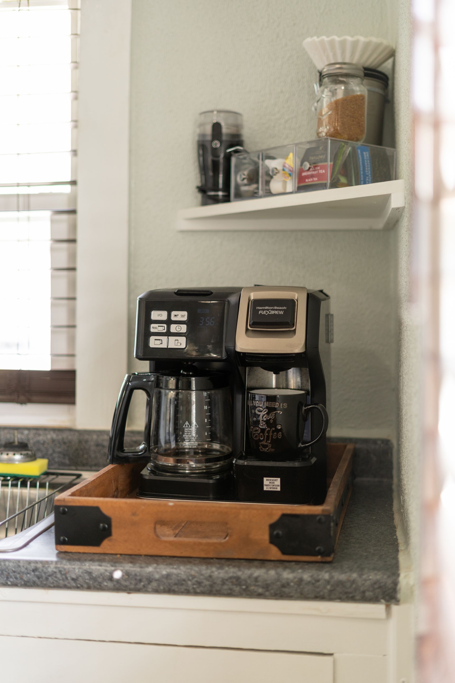 Coffee maker on a tray in a kitchen; shelf above holds coffee filters and supplies.