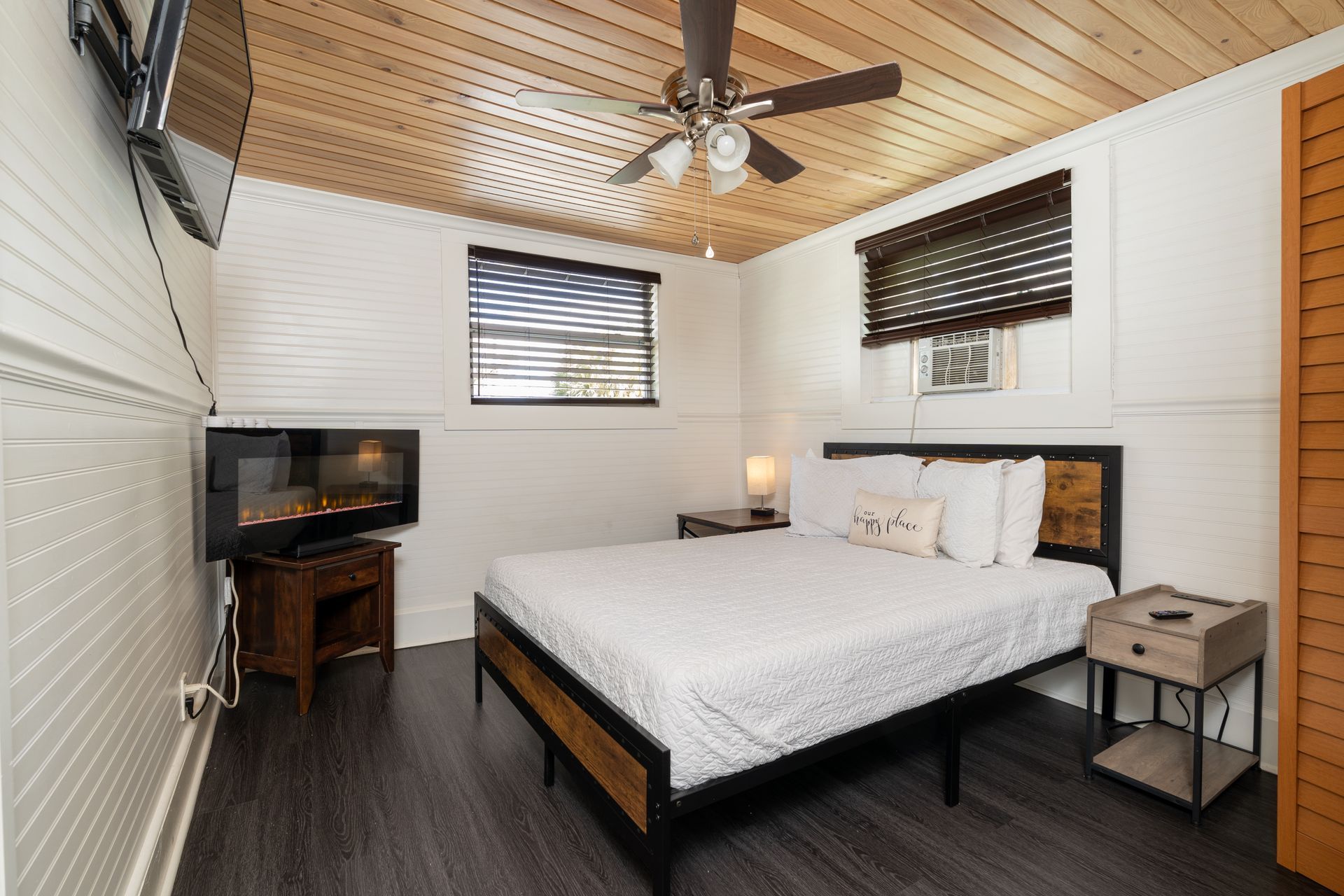 Bedroom with a bed, fireplace, TV, ceiling fan, wood paneling, and a window.