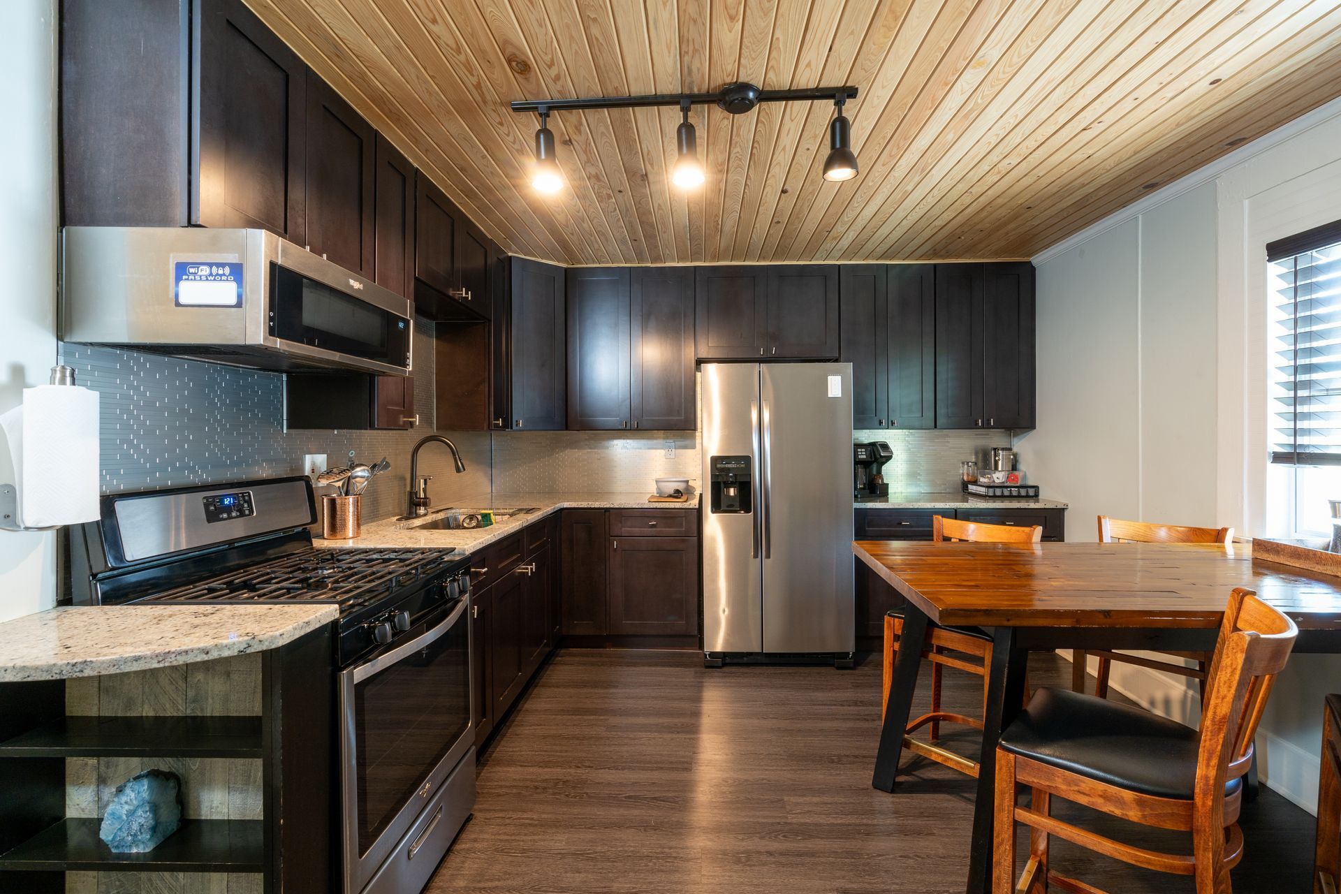 Modern kitchen with dark cabinetry, stainless steel appliances, and wood ceiling. Includes a dining table.