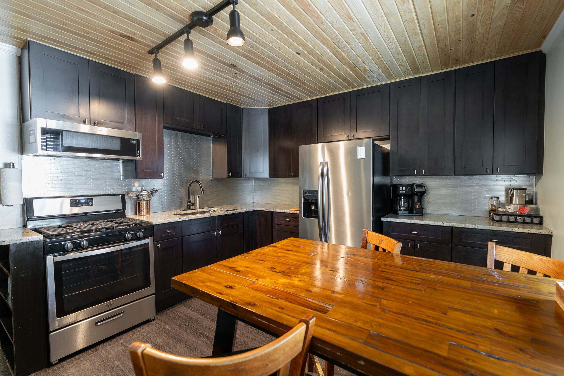 Kitchen with dark cabinets, stainless steel appliances, wood table, and ceiling.
