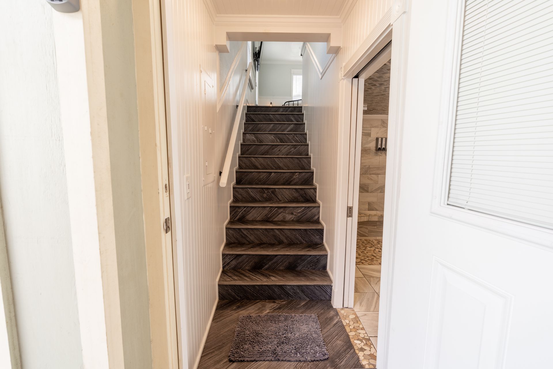 Narrow hallway with stairs leading up, door on the right, light-colored walls, dark stair treads.