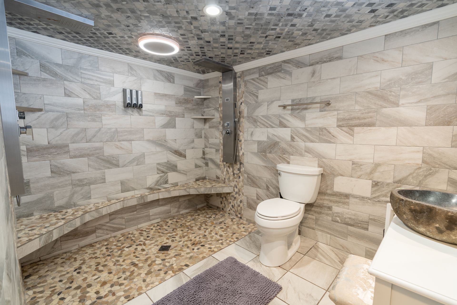 Bathroom with stone tile shower and pebble floor, toilet, vanity with sink, and gray rug.