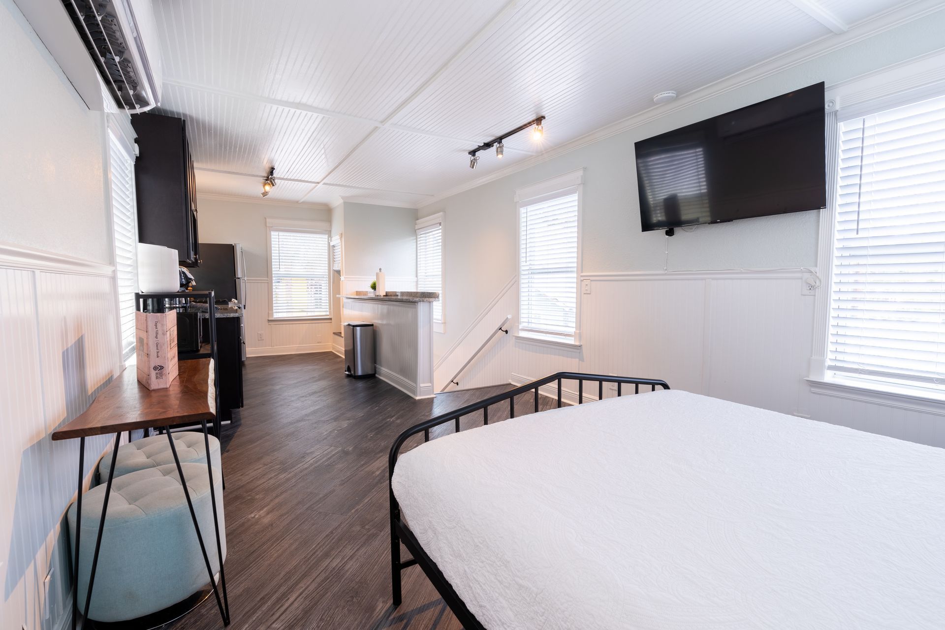 Small studio apartment with bed, TV, kitchen, and desk. Bright white walls, dark floor.