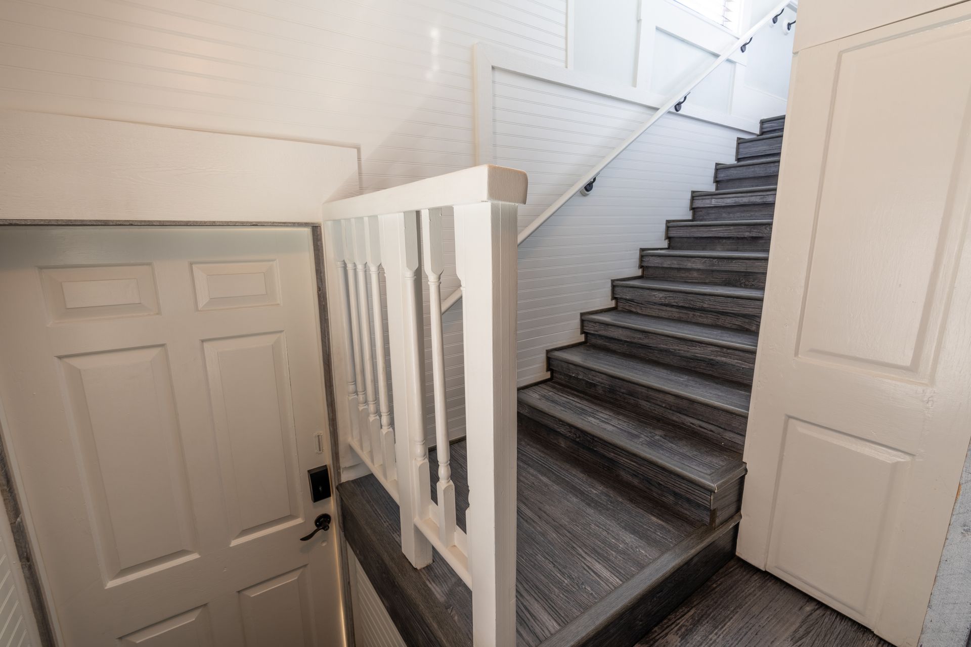 Staircase with gray wooden steps, white banister and door, and white walls.
