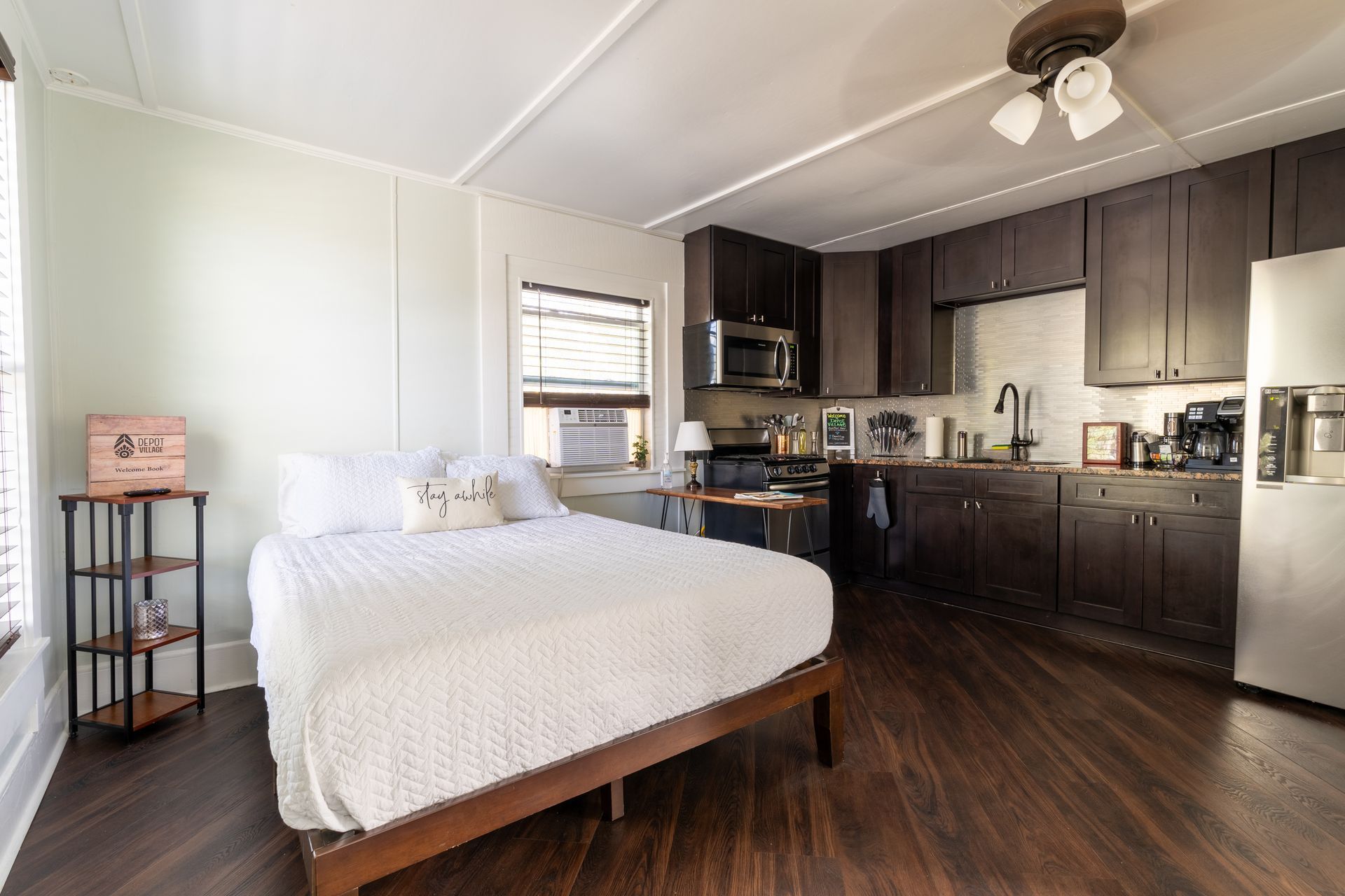 Small studio apartment with bed, dark cabinets, and stainless steel fridge. Wooden floor.