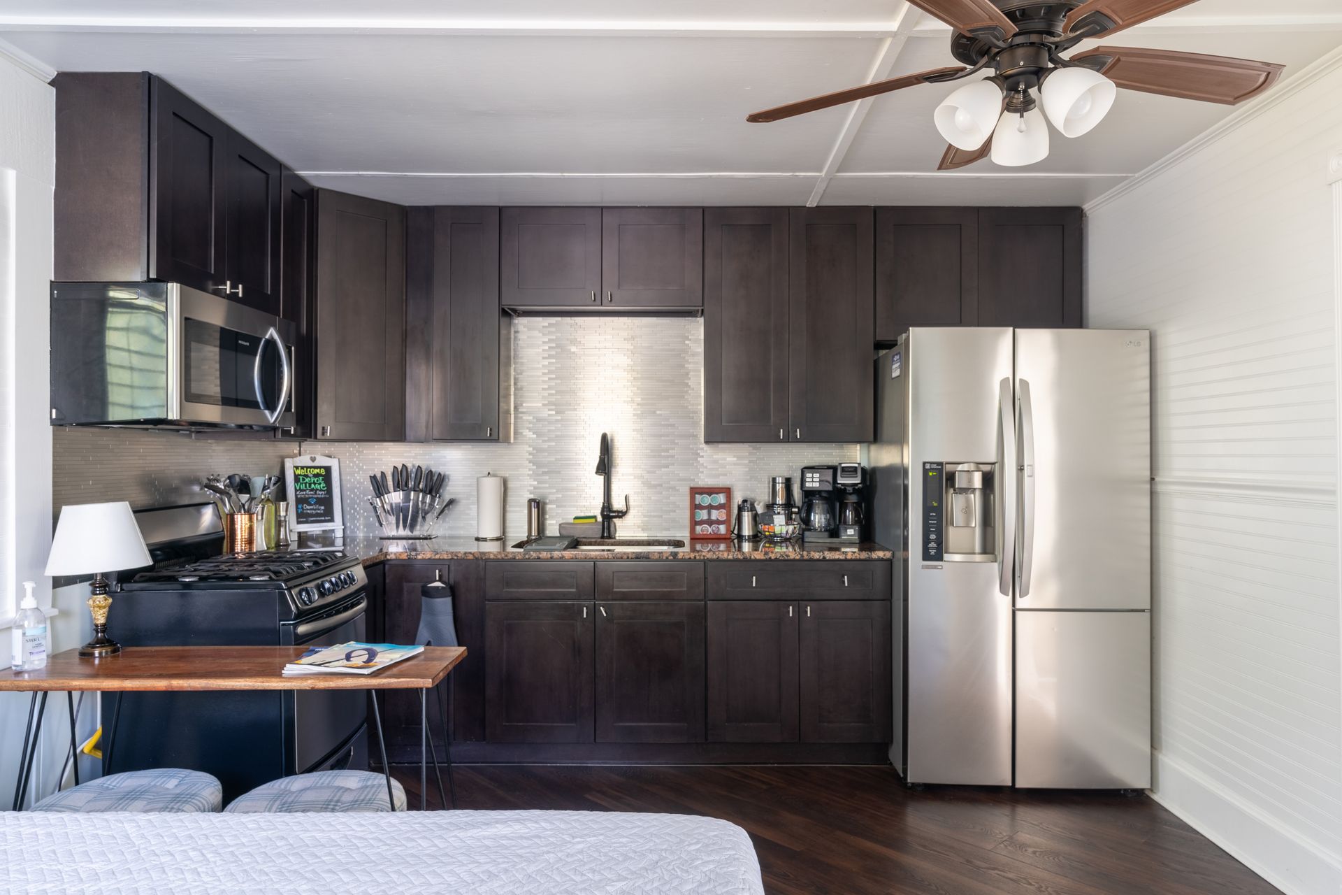 Dark-cabinet kitchen with stainless appliances. Includes a stove, refrigerator, and a table. Brown ceiling fan above.