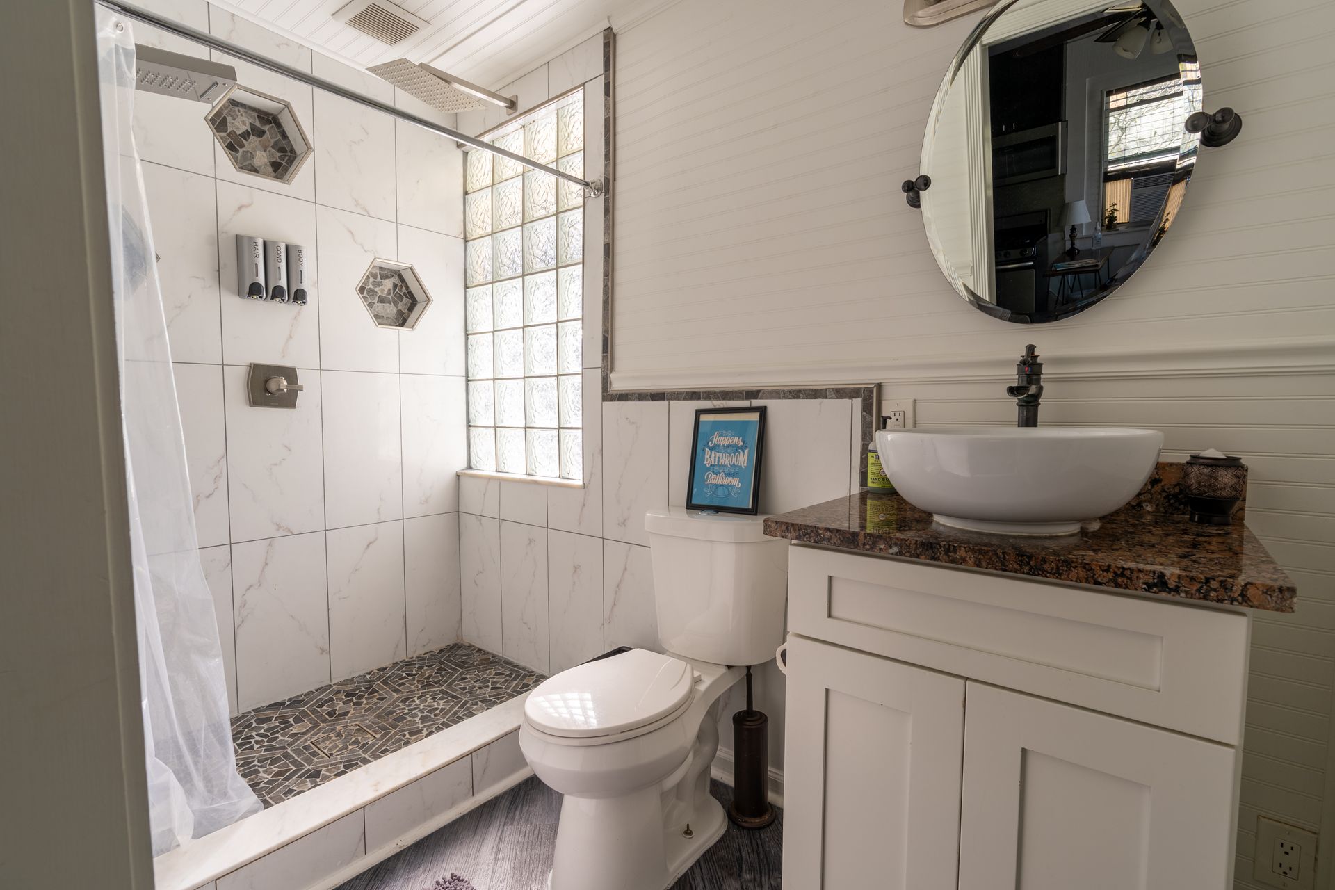 Bathroom with a white toilet, white and grey tiled shower, vanity with a vessel sink and oval mirror.