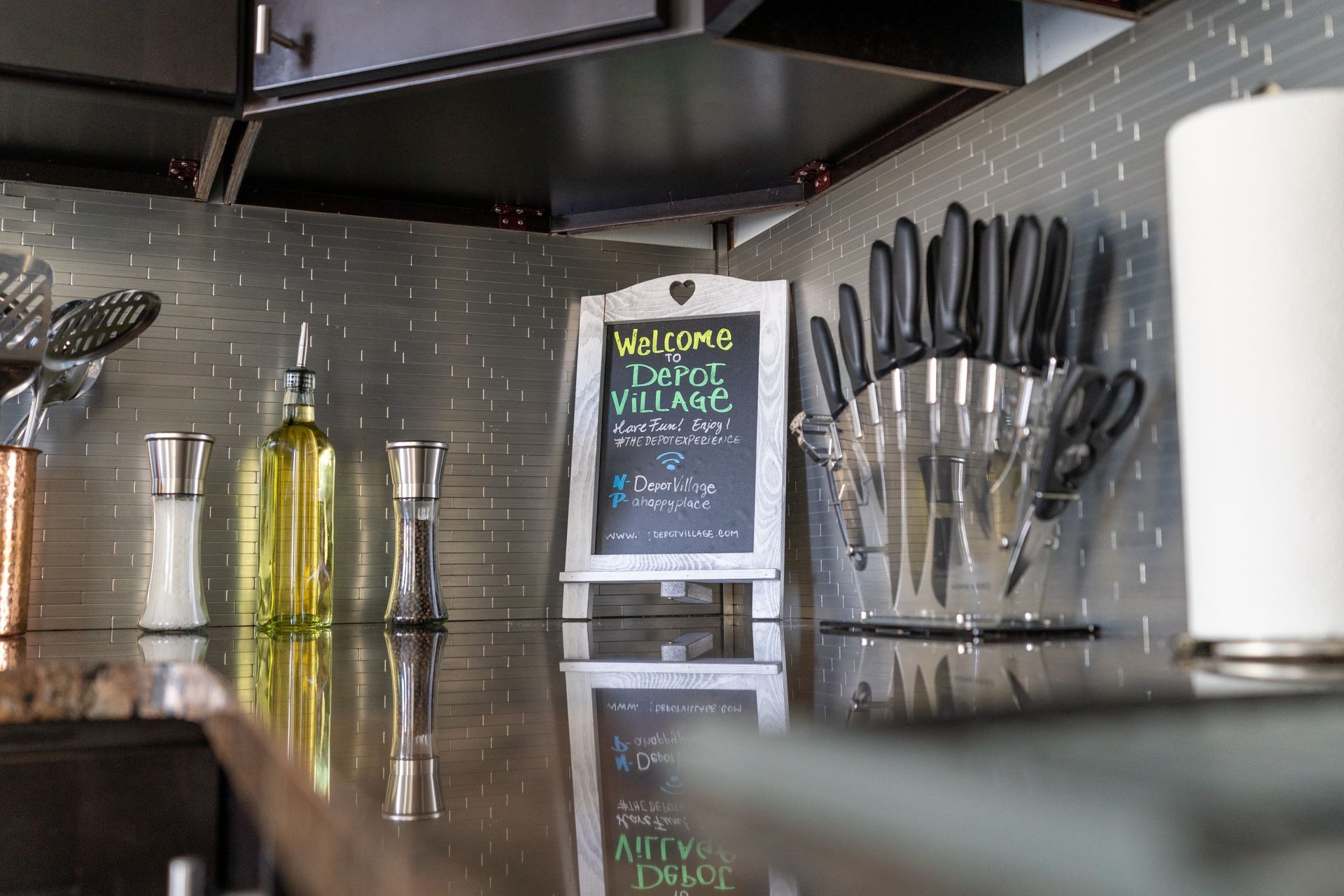 Kitchen counter with utensils, oil, spices, knife block, and welcome sign.