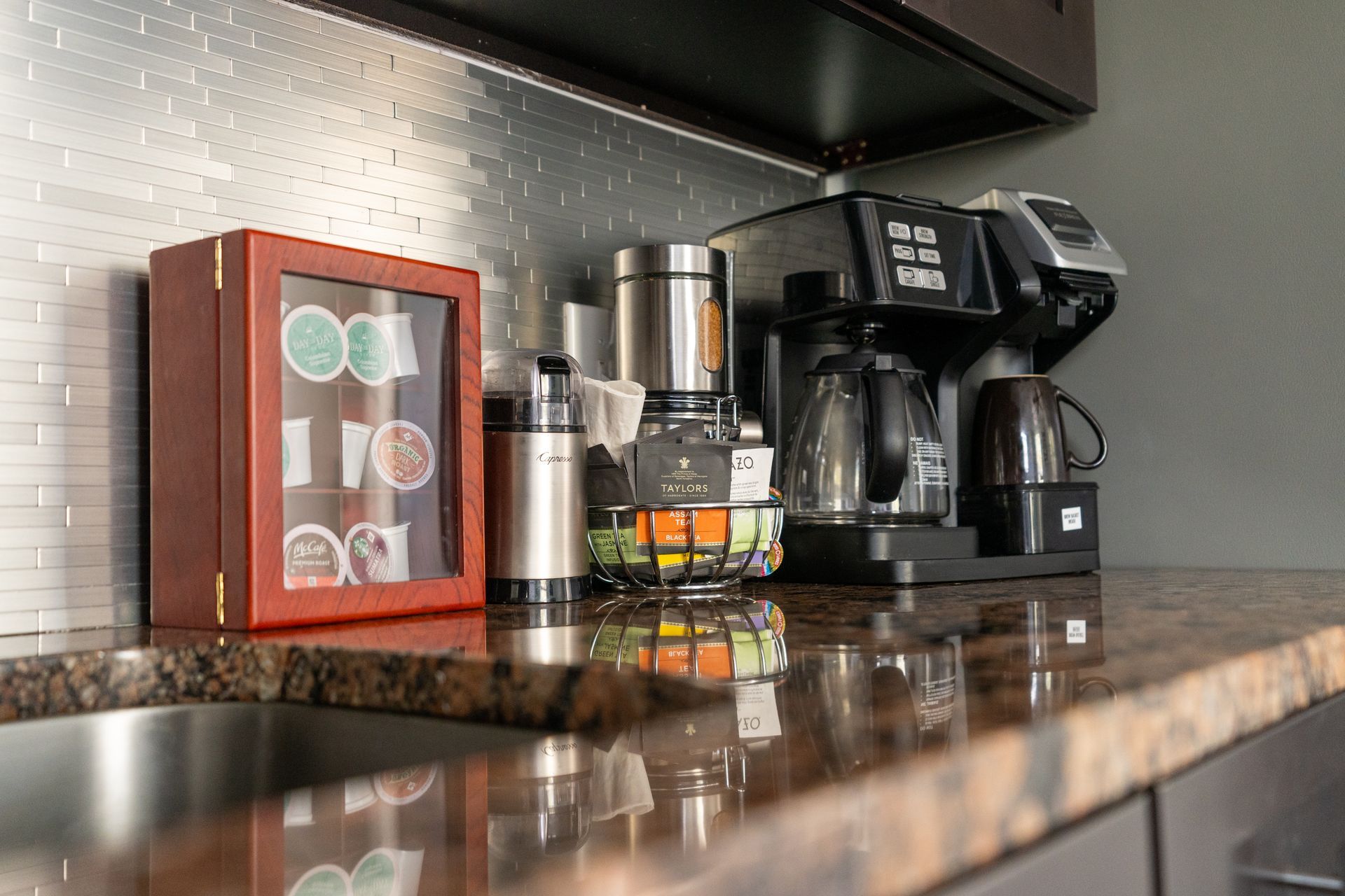 Coffee station: Keurig machine, coffee pods, water kettle, fruit basket, and stainless steel thermos on granite countertop.