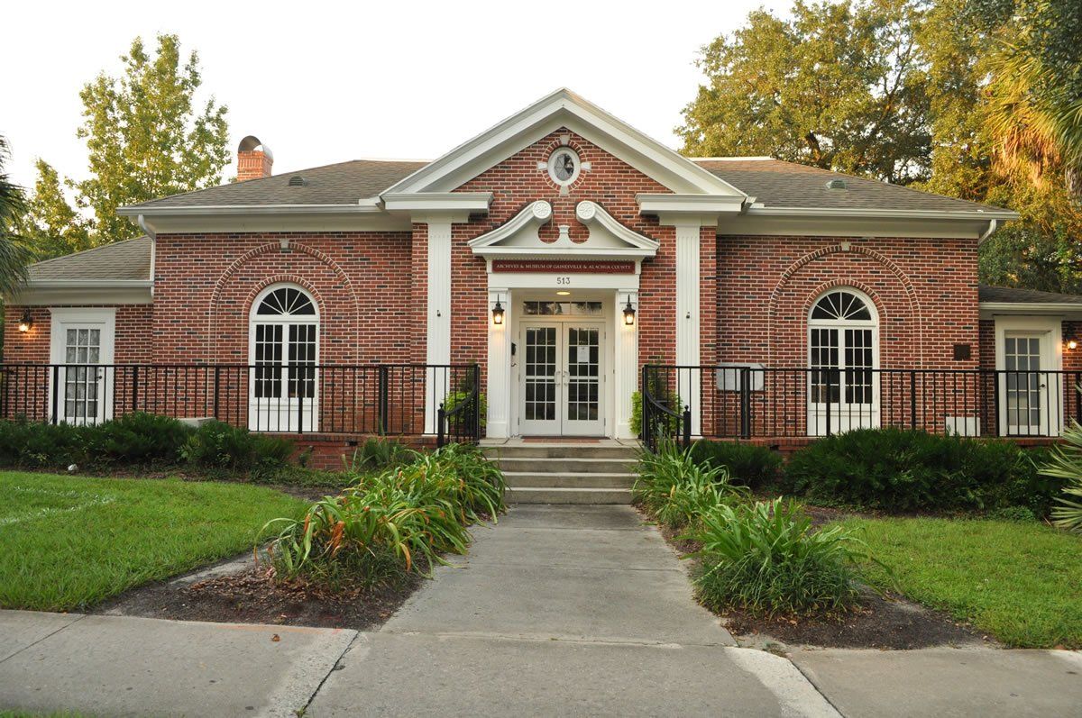 Brick building with white trim, arched windows, and double doors; walkway leads to entrance, flanked by grass and shrubs.