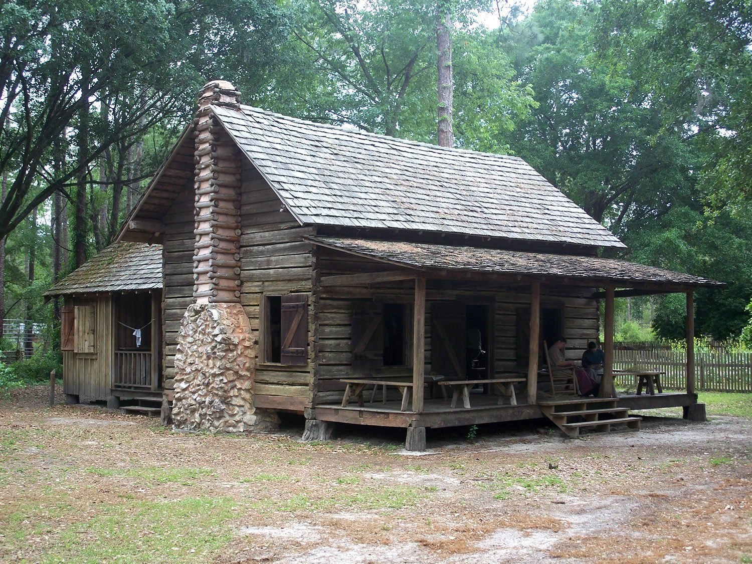 Wooden log cabin with stone chimney and porch, set in a wooded area.