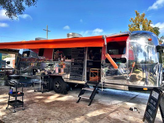 Shiny, silver food truck with orange awning, open serving window, parked outdoors under blue sky.