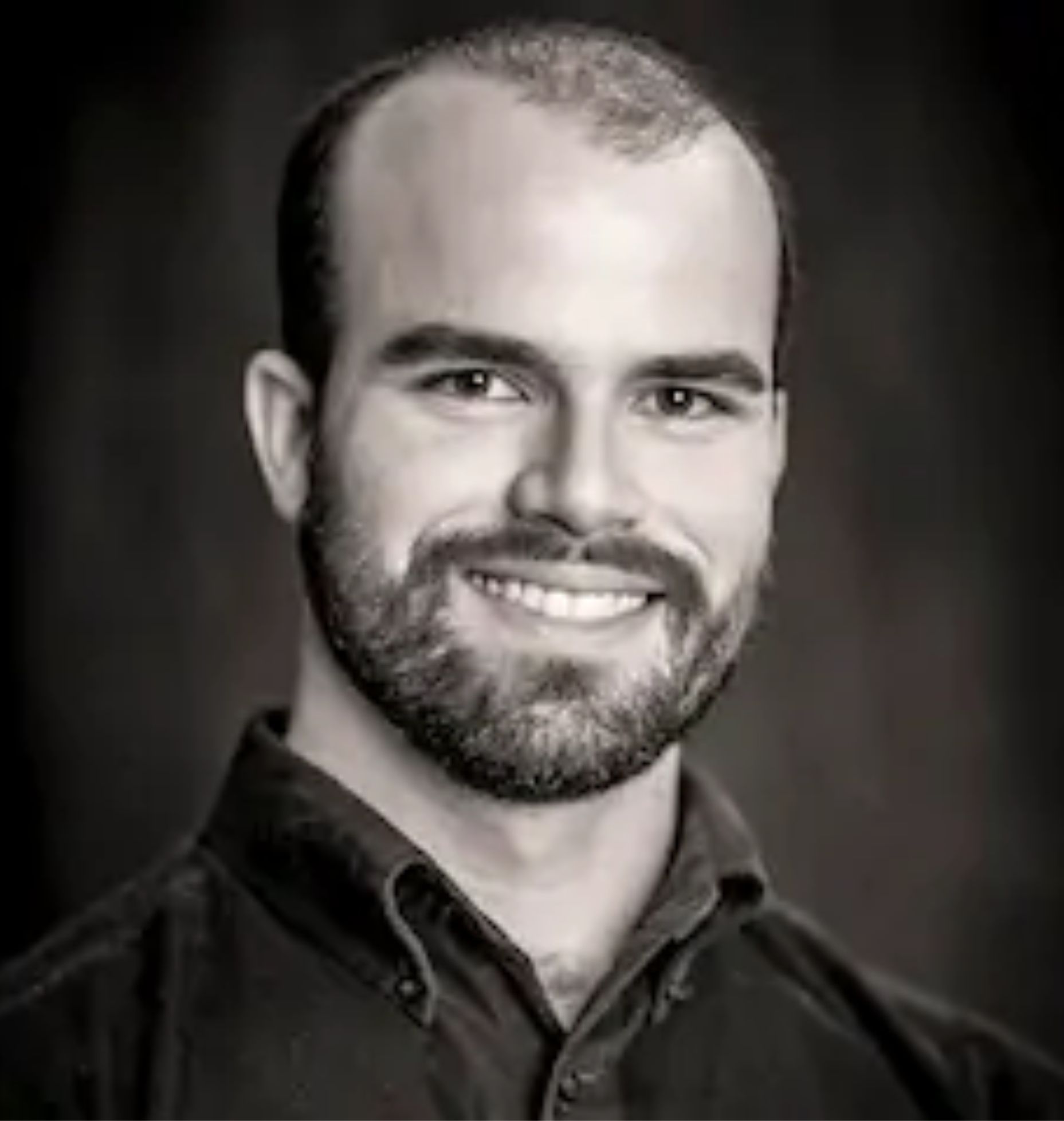 Man with a beard, smiling, wearing a dark button-up shirt, against a dark blurred background.