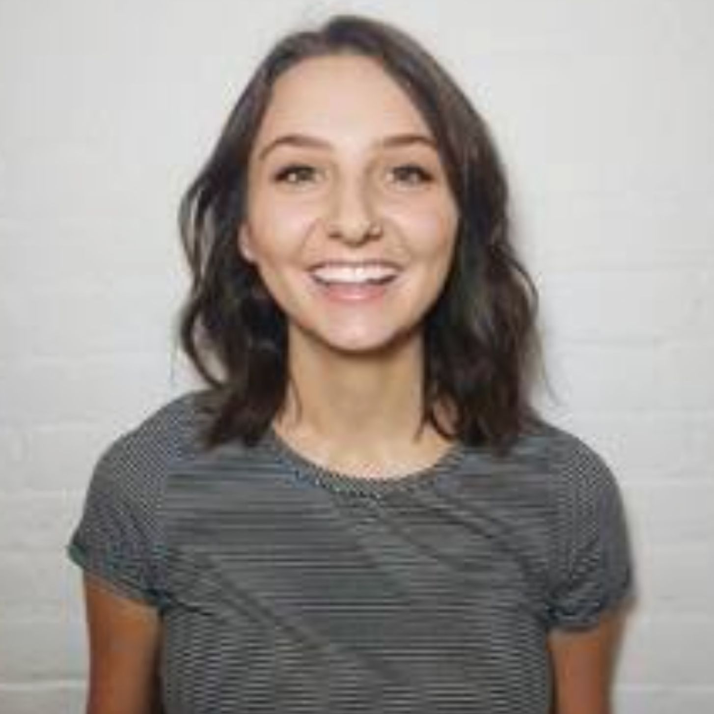 Woman smiling, wearing a striped shirt, standing against a white wall.