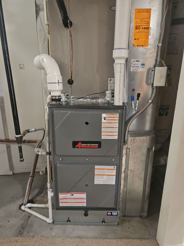 Gray furnace unit with white PVC vent pipes and orange warning labels in a basement utility room