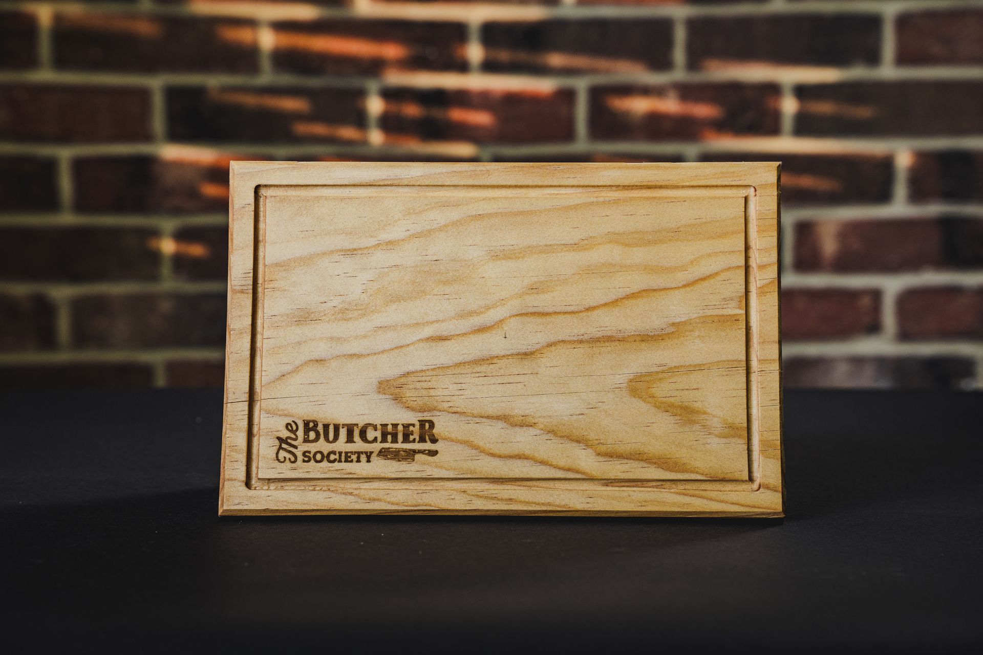 Tabla de picar “The Butcher Society”, apoyada contra un árbol al aire libre sobre el césped.